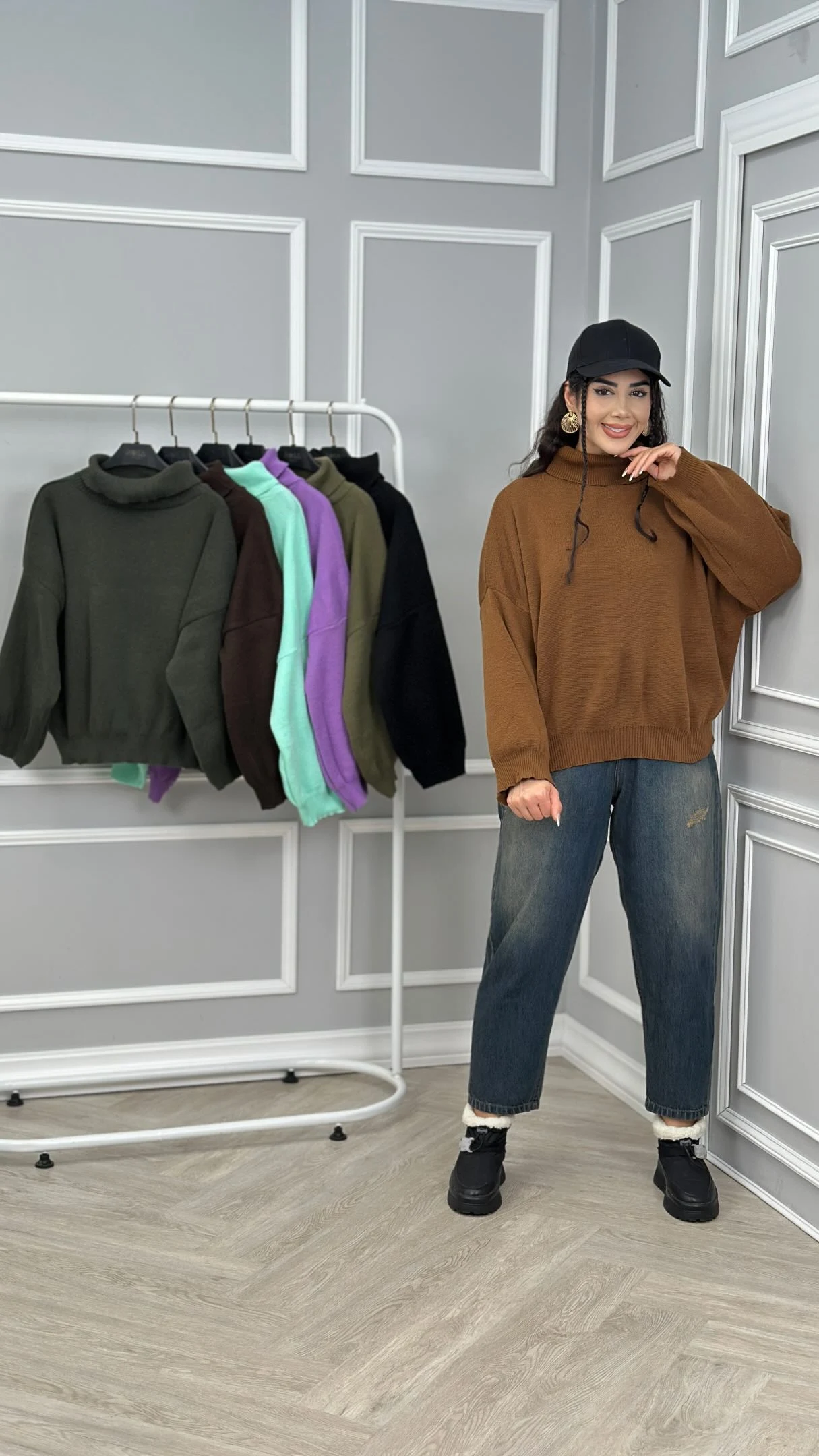 Model in a loose brown turtleneck, cuffed blue jeans, black cap, and black lace-up shoes.