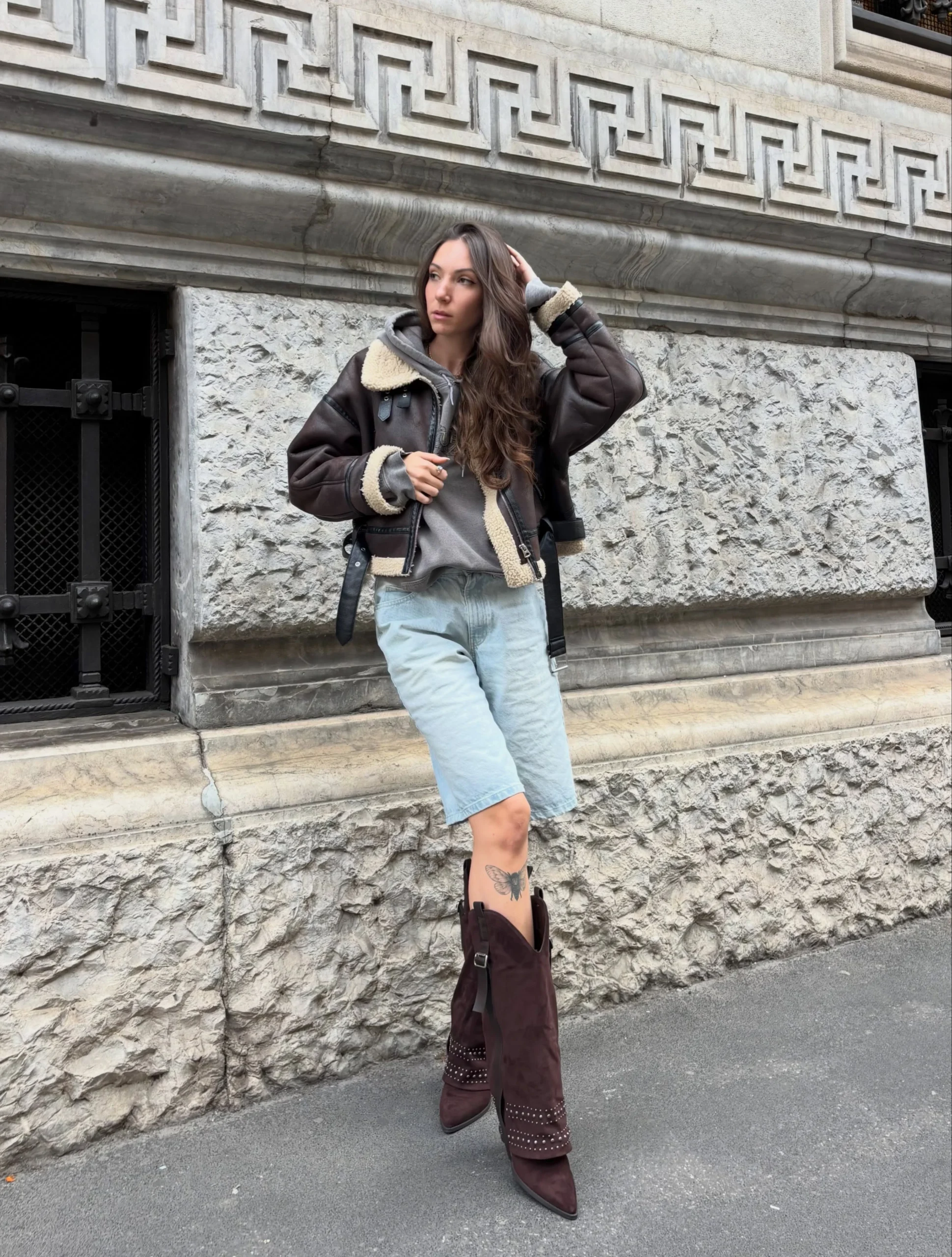 Dark brown shearling jacket worn open over gray hoodie, light-wash denim shorts, and tall studded boots.