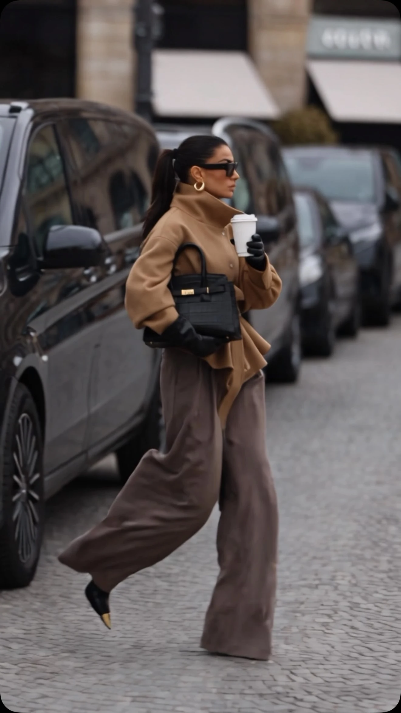 Person in an open camel coat, wide dark brown trousers, and black pointed shoes on a city street.