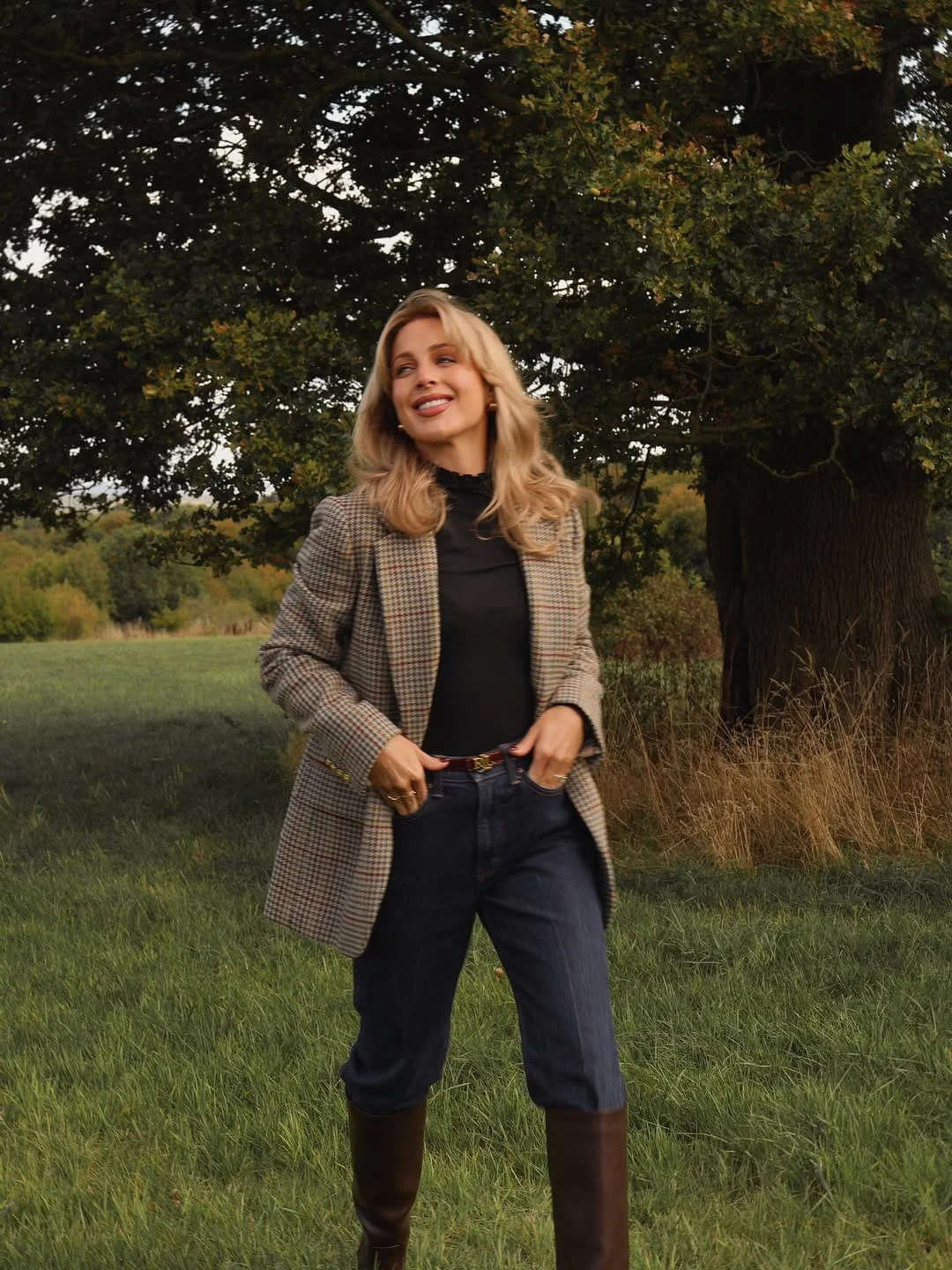 A checked blazer worn over a black ruffle-neck top, paired with straight-leg dark-wash jeans and tall brown riding boots, is the country chic look at its most versatile — the dark denim and black top create a strong, unfussy base that lets the blazer pattern do its work without competition, and the riding boots are the piece that keeps it in countryside territory rather than slipping into city smart-casual. The ruffle neck is the detail that elevates this above a straightforward blazer-and-jeans formula: it adds a softness and femininity at the neckline that makes the checked blazer feel more special and considered. I find this completely irresistible for the transition from a countryside walk straight to a cosy pub — it works equally well in both contexts.