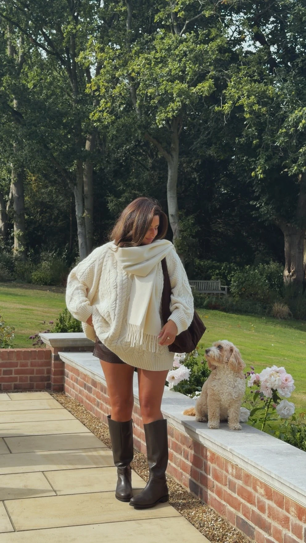 Oversized cream cable-knit sweater with matching fringed scarf, dark shorts, tall brown boots, and shoulder bag.