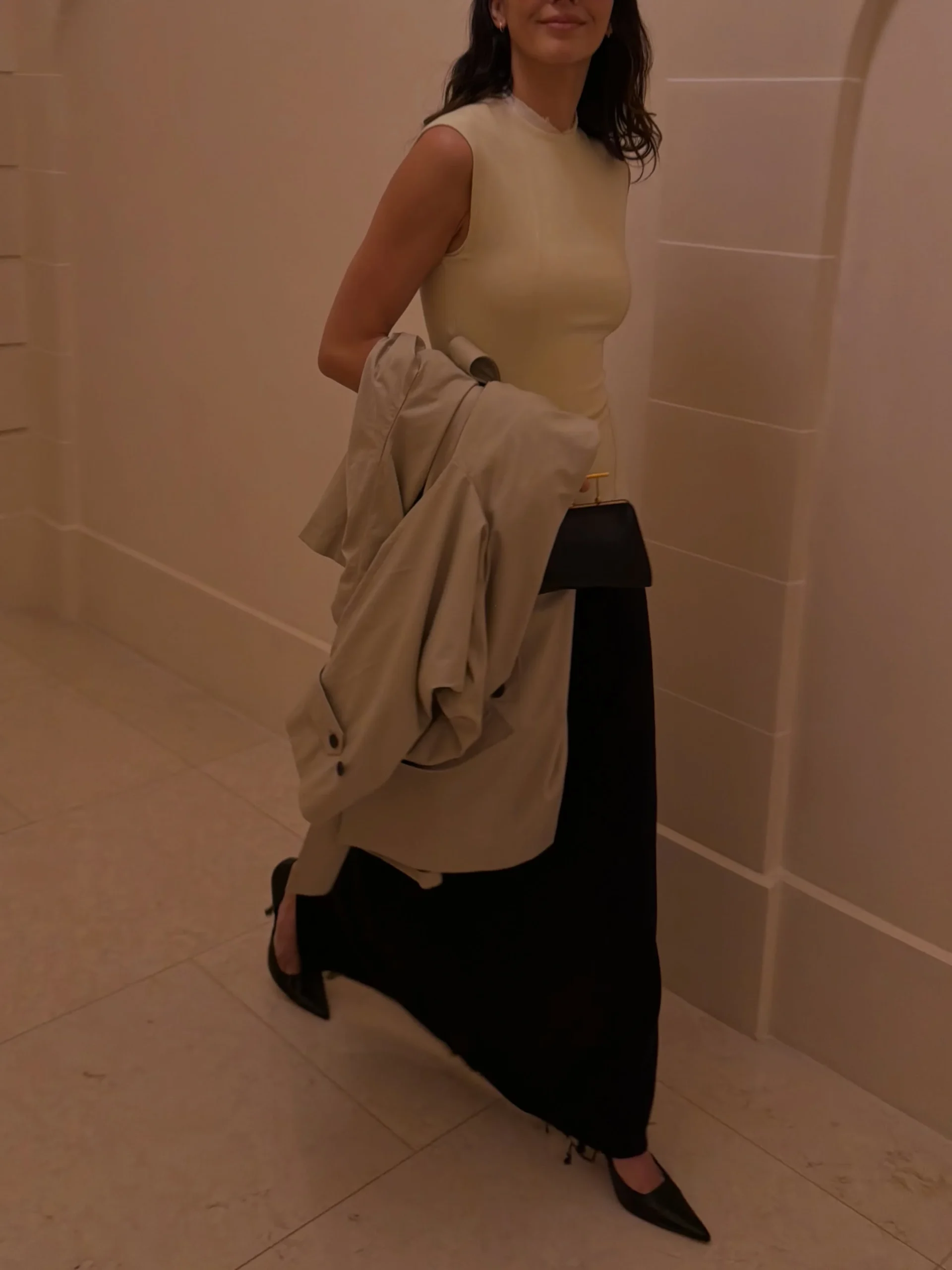 Person in cream sleeveless top, long black skirt, and black pointed-toe shoes carrying a beige jacket.