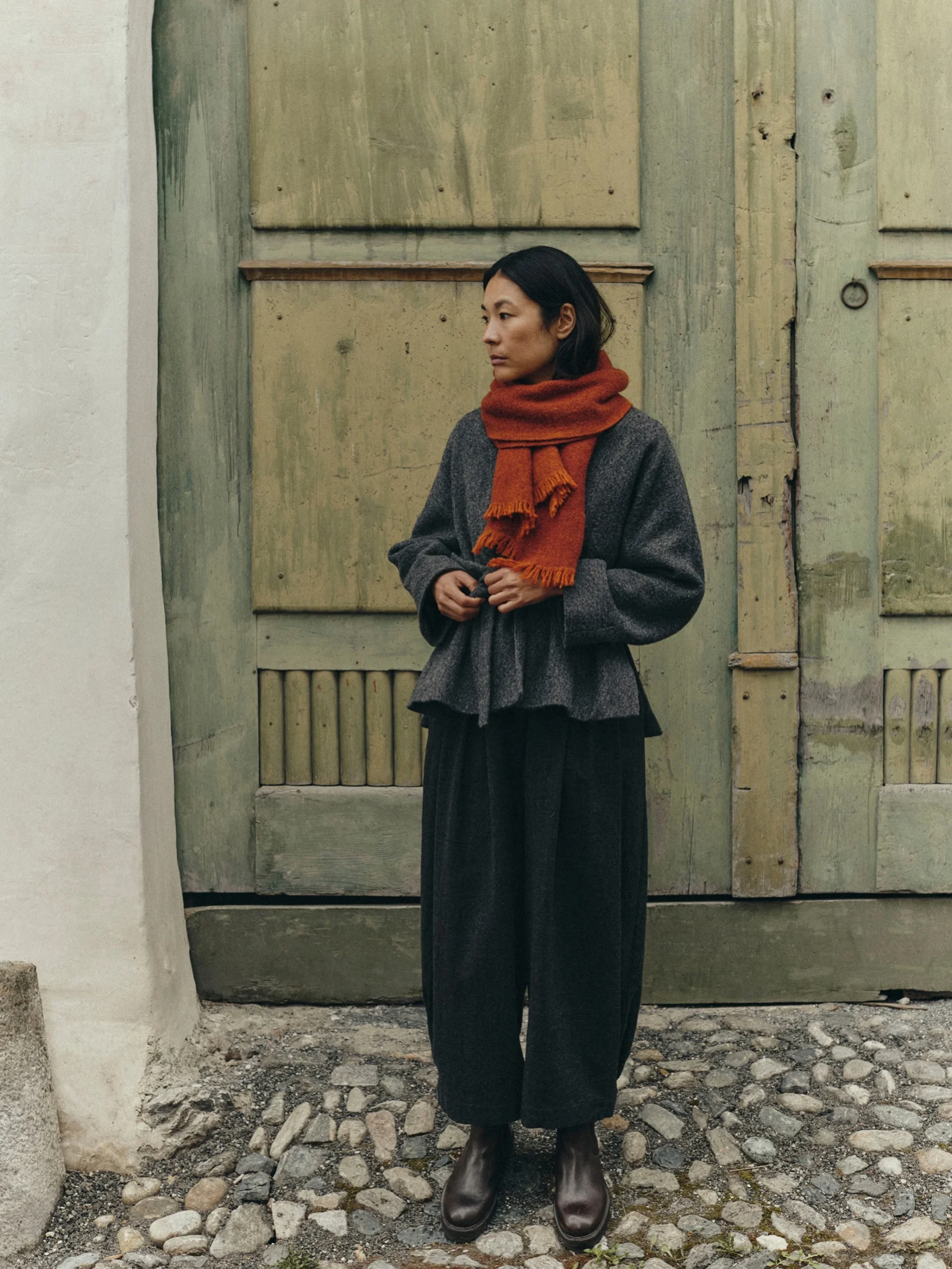 Dark gray loose wool coat, wide-leg black trousers, dark brown leather boots, bright rust-orange fringed scarf