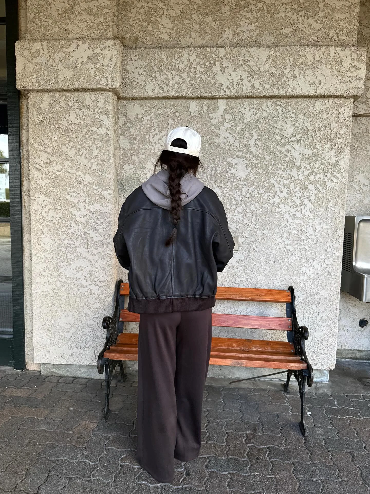 Grey hoodie under black leather bomber with wide-leg brown trousers, white sneakers, and backward cap.