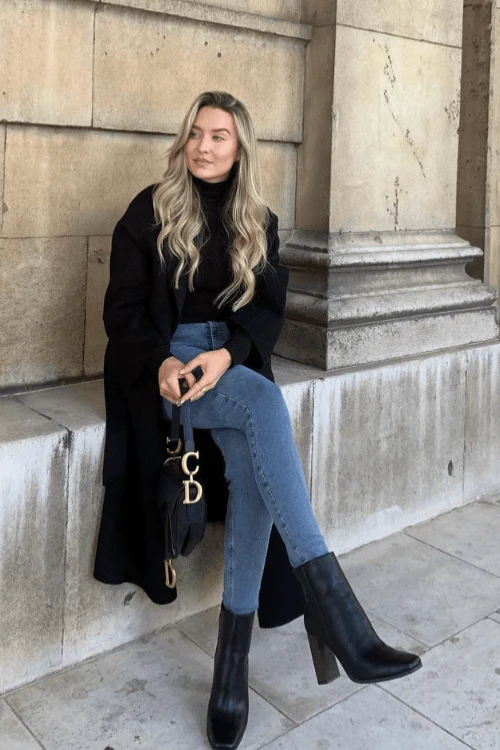 Woman in long black duster coat, fitted turtleneck, skinny jeans, and black block-heel ankle boots.