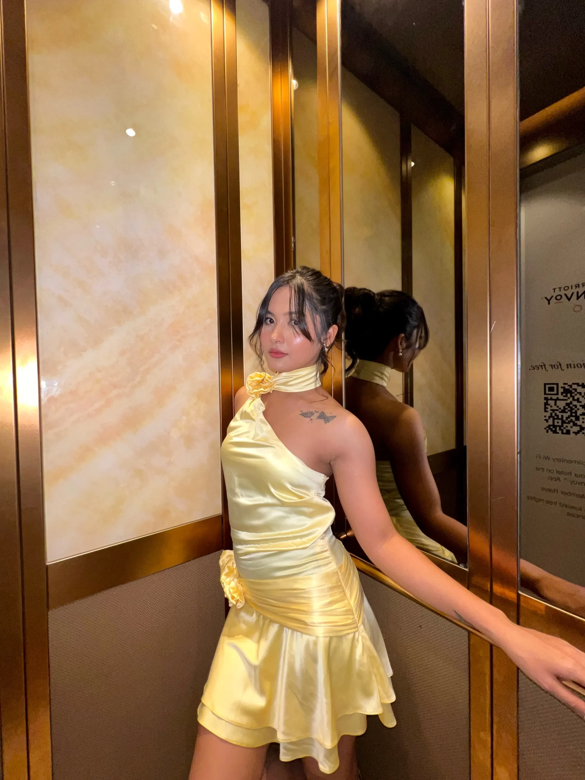 Woman wearing a pale-yellow satin one-shoulder mini dress with floral accents and pearl earrings.