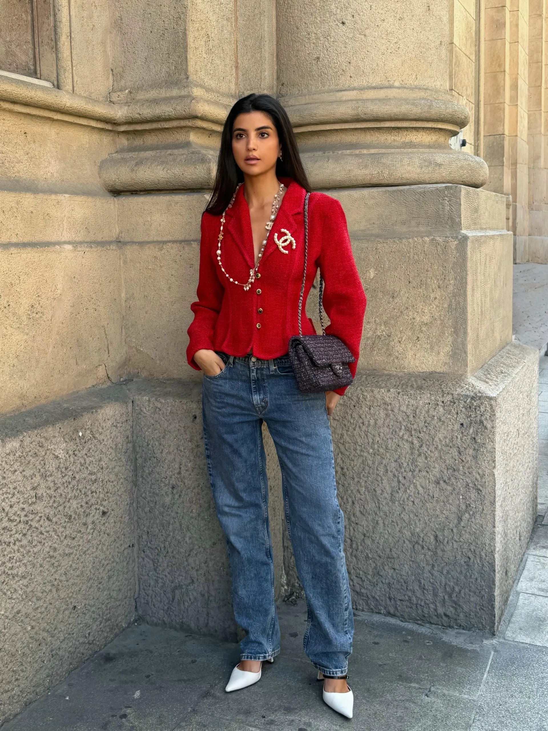 Person wearing a bright red V-neck cardigan with blue jeans, white shoes, and a quilted bag outdoors.