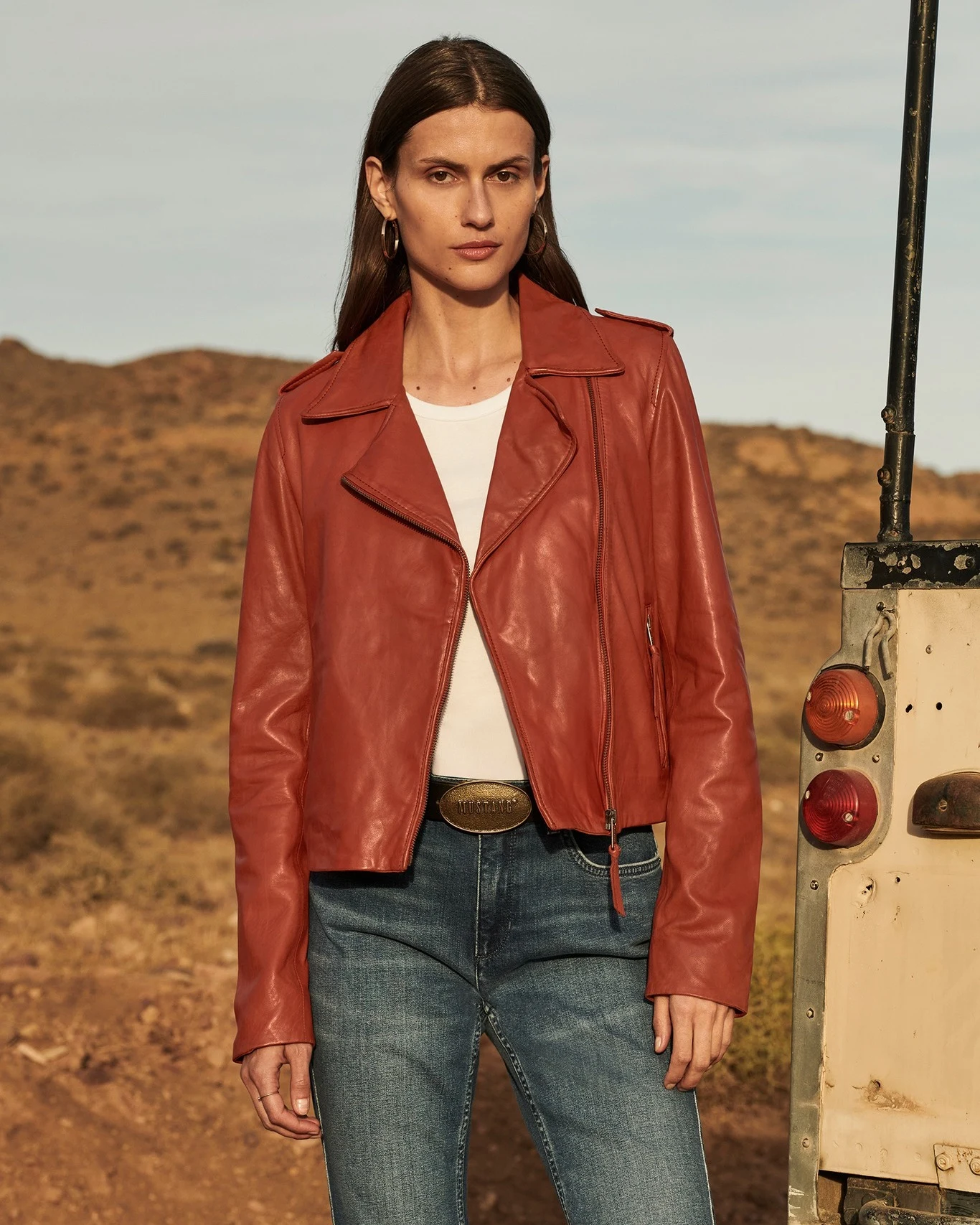 rust-red leather moto jacket with asymmetrical zip and shoulder epaulets, plain white crew-neck t-shirt, medium-wash blue straight-leg denim jeans, dark belt with oval brass buckle, gold hoop earrings