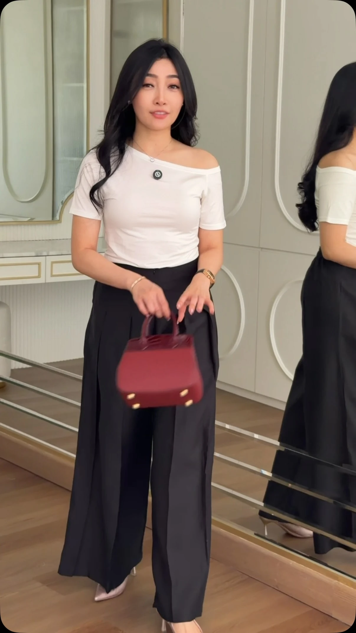 Model in a white off-shoulder top and black pleated wide-leg trousers holding a burgundy handbag.