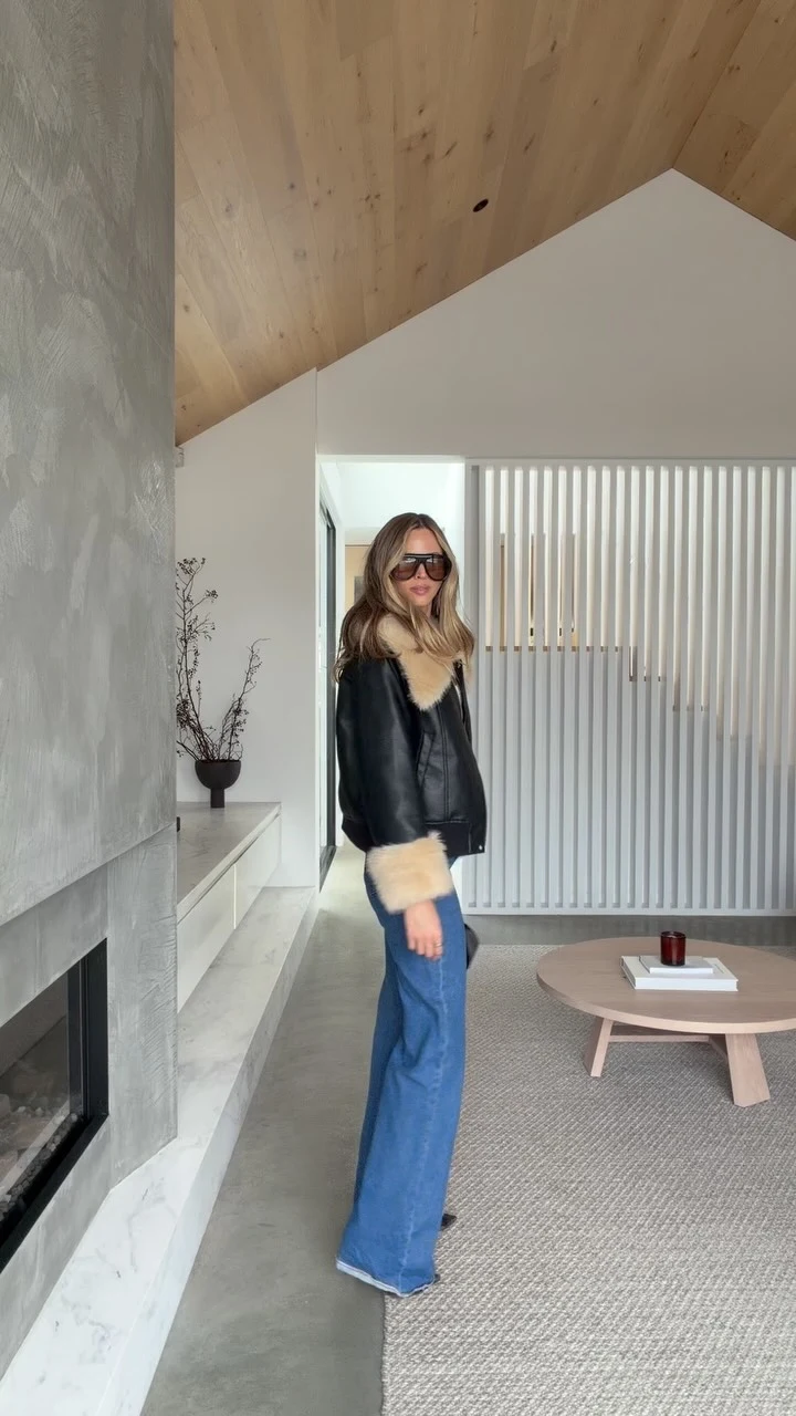 Black shearling jacket with cream collar and cuffs styled with floor-length blue wide-leg jeans and dark sunglasses.