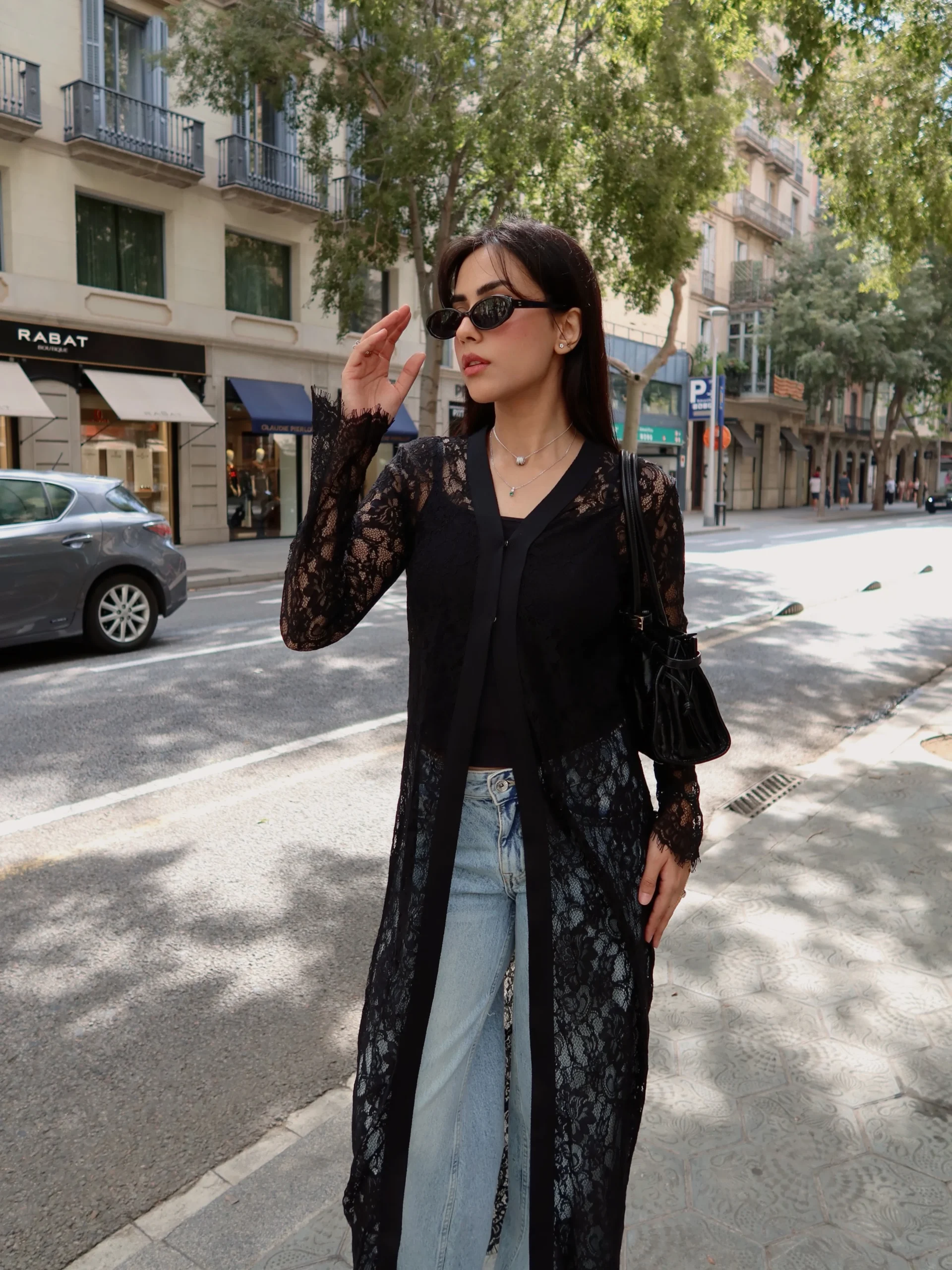 Black lace duster worn open over a black top with light-wash jeans, shoulder bag, and sunglasses.