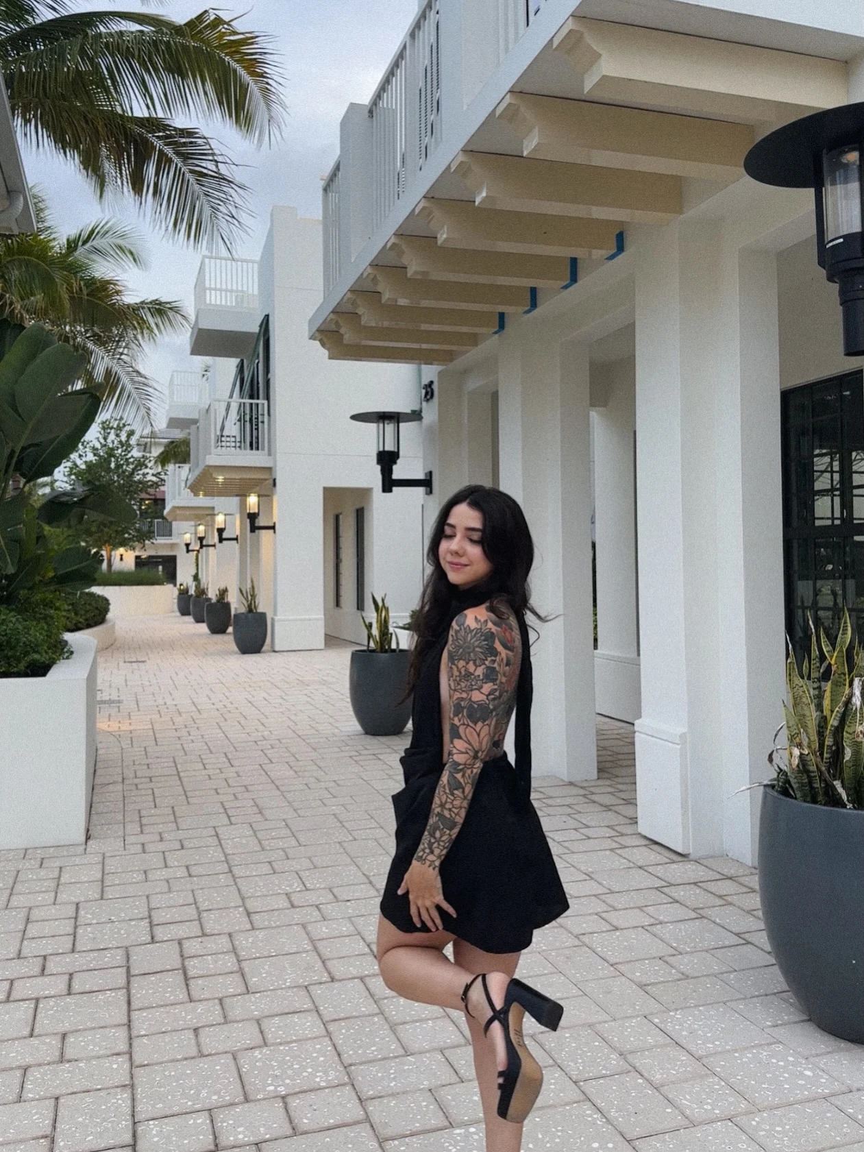 Woman in a black sleeveless flared mini dress and black block-heel sandals on an outdoor walkway.