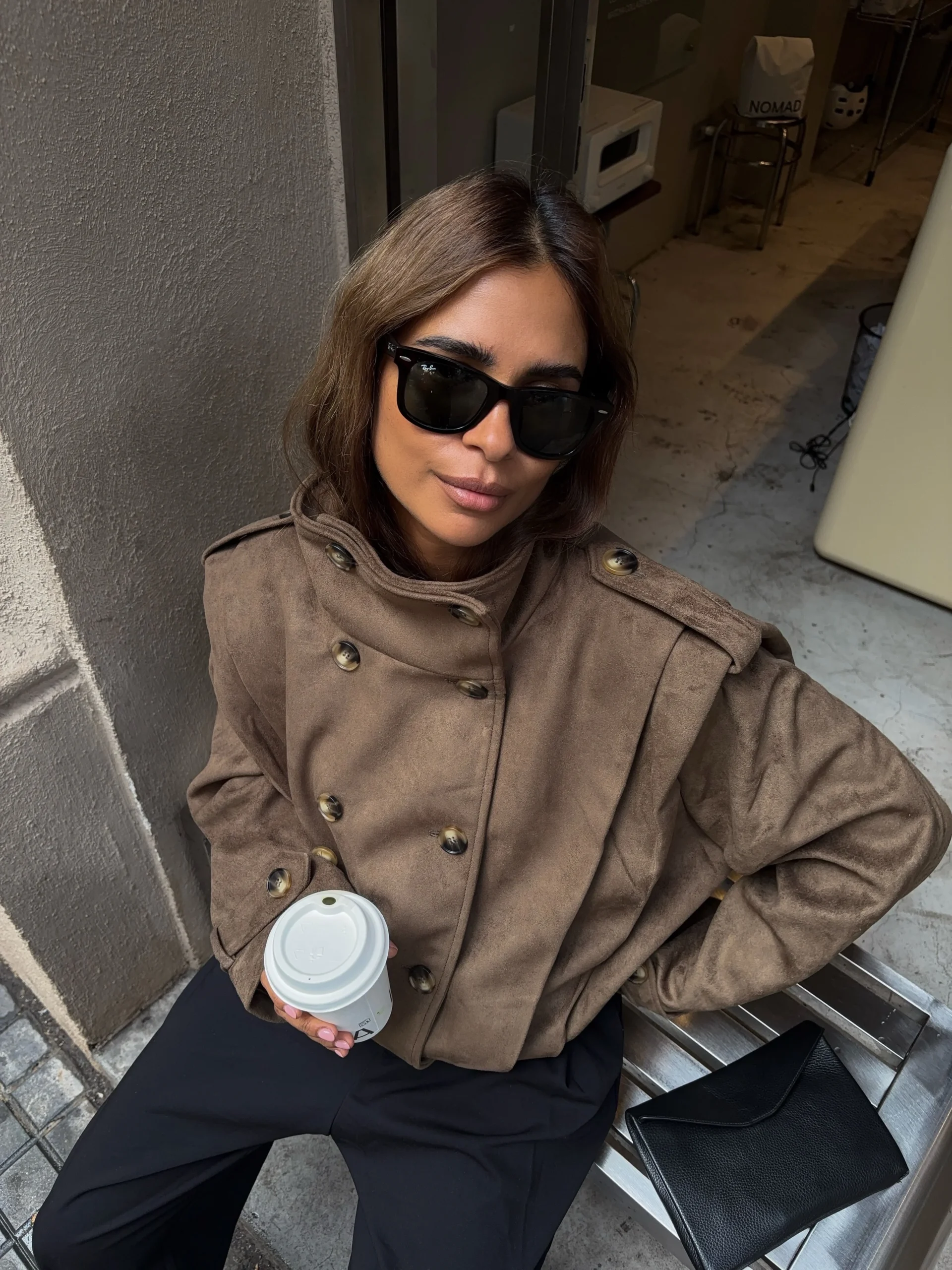 Brown waist-length structured jacket with dark buttons worn with black trousers, sunglasses, and a rectangular bag.