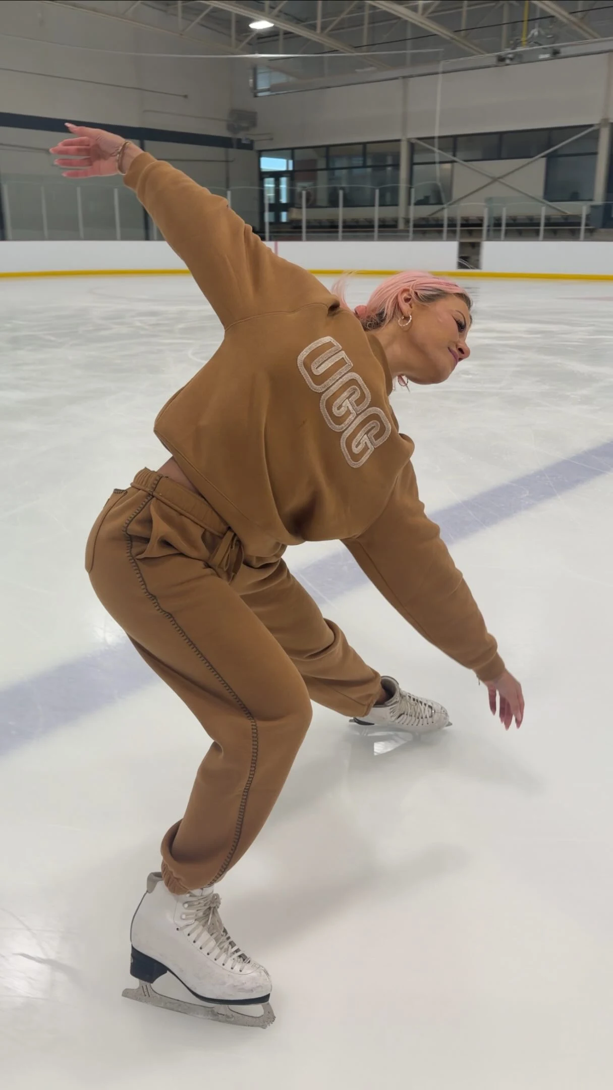 Skater in camel sweatshirt and matching brown pants with white figure skates, leaning sideways on an ice rink.