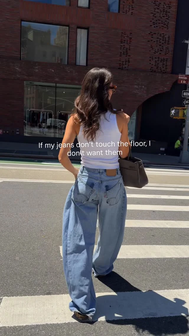 Woman in floor-length baggy light-blue jeans, white crop tank, tan slip-ons, and brown shoulder bag.