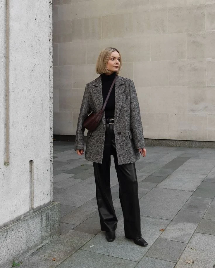 Woman in grey tweed blazer, faux leather trousers, black turtleneck, and pointed ankle boots with crossbody bag.
