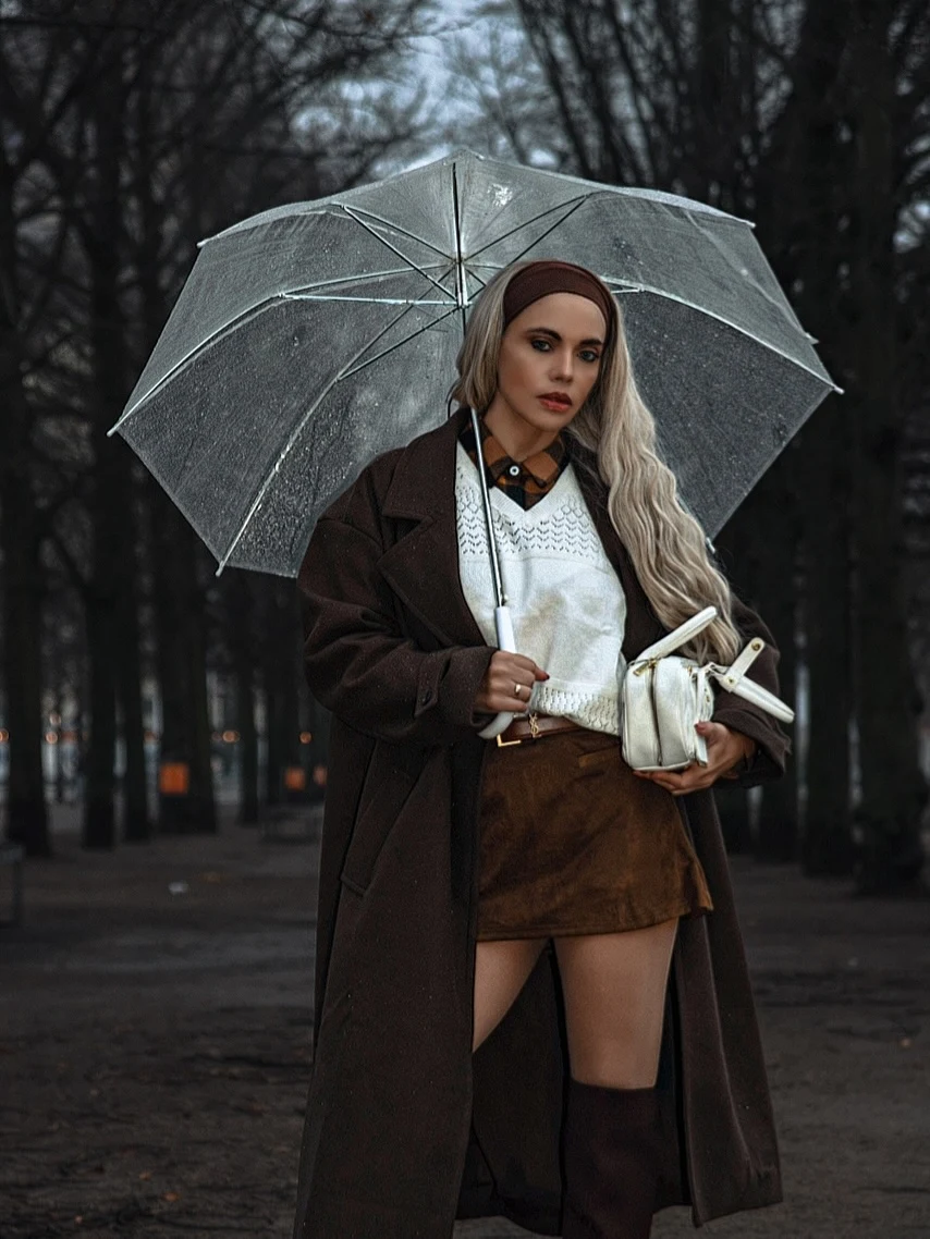 Long dark brown coat over white V-neck sweater and short brown skirt, knee-high socks, brown headband, transparent umbrella overhead.