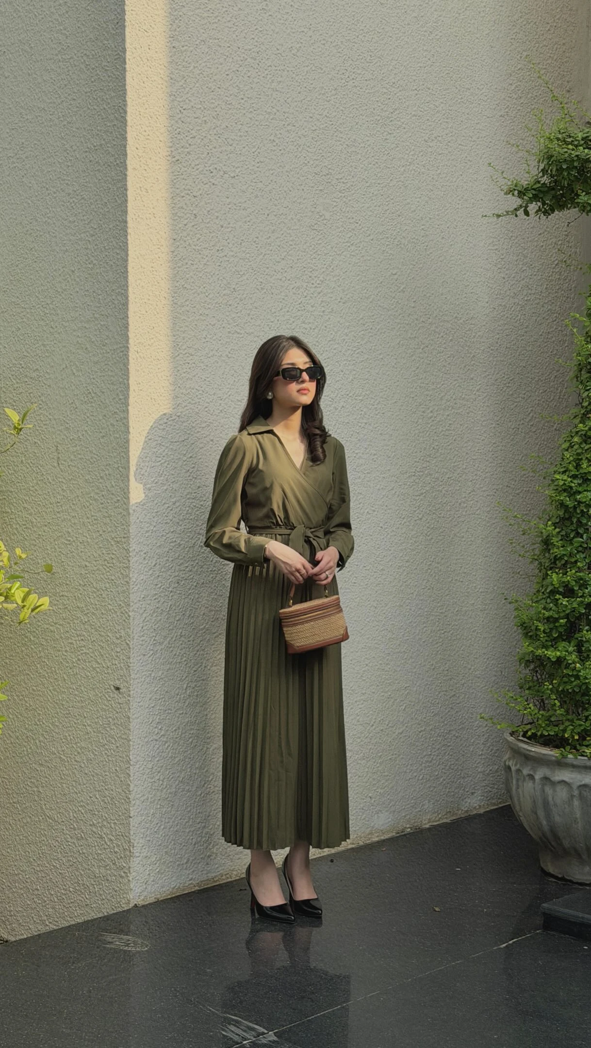 Long olive green wrap dress with pleated skirt, waist belt, black shoes, and a small tan bag.