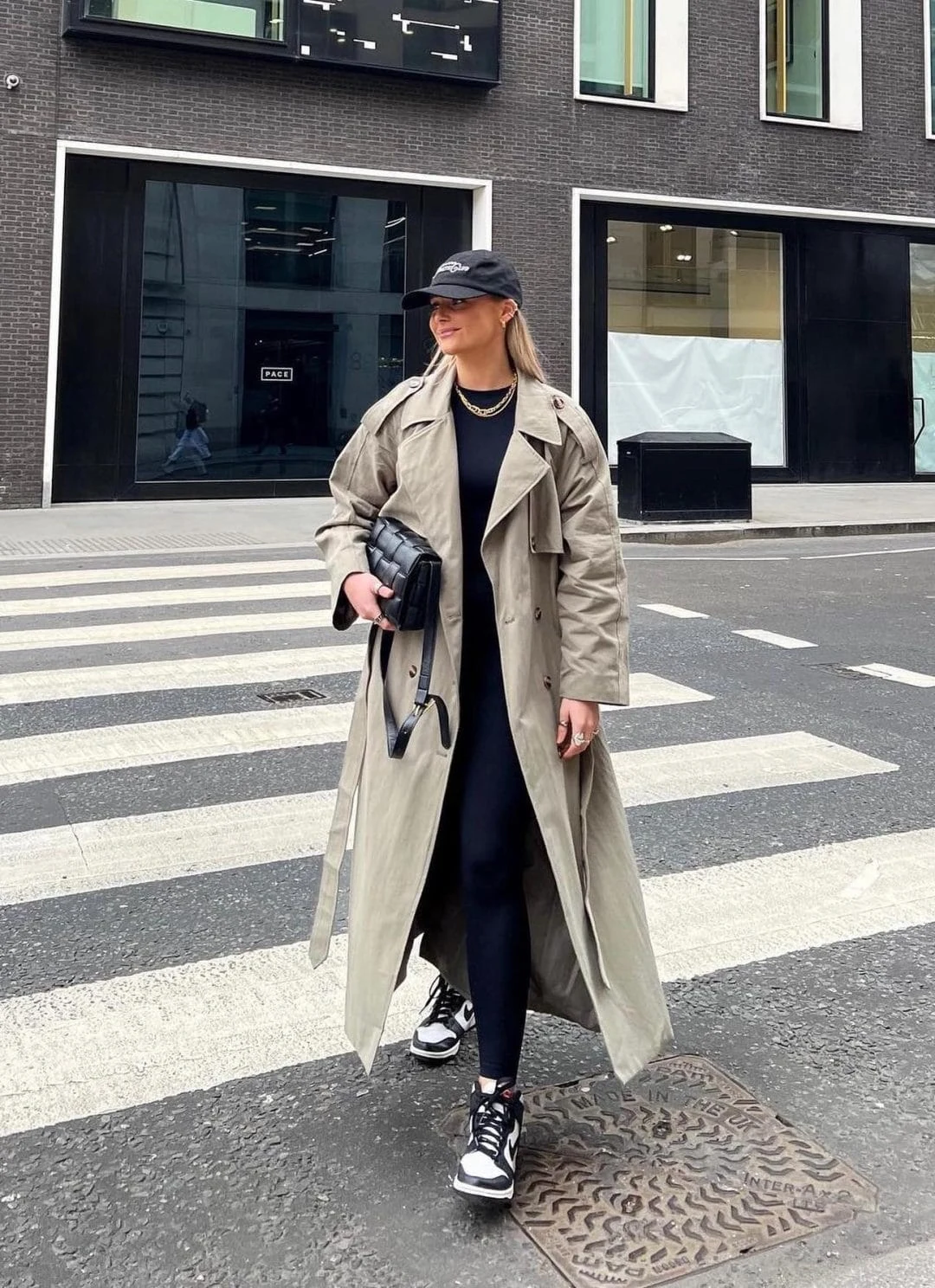 This outfit blends sporty and chic with an oversized khaki trench coat over a sleek black leggings and top set. A black cap, layered gold necklaces, a quilted clutch bag, and black-and-white sneakers complete this effortlessly stylish, laid-back city look.