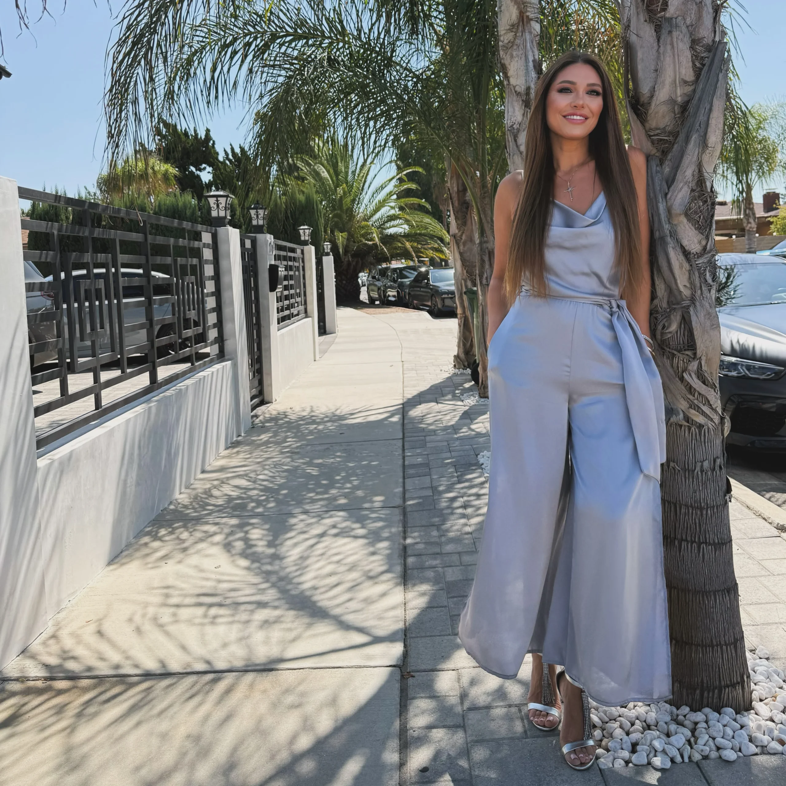 A pale blue sleeveless jumpsuit has a draped cowl neckline, a fitted waist with a tied sash, and wide legs that fall to the ankles. The woman wears silver-toned open-toe heels. The outfit is shown outdoors beside a palm tree on a sunny sidewalk.