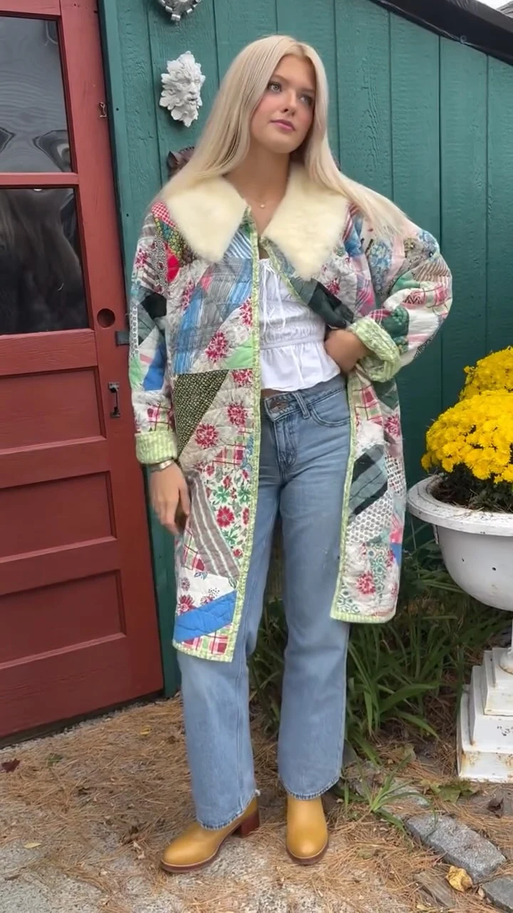 Person in a colorful patchwork quilted coat, white top, blue jeans, and mustard ankle boots by a red door.