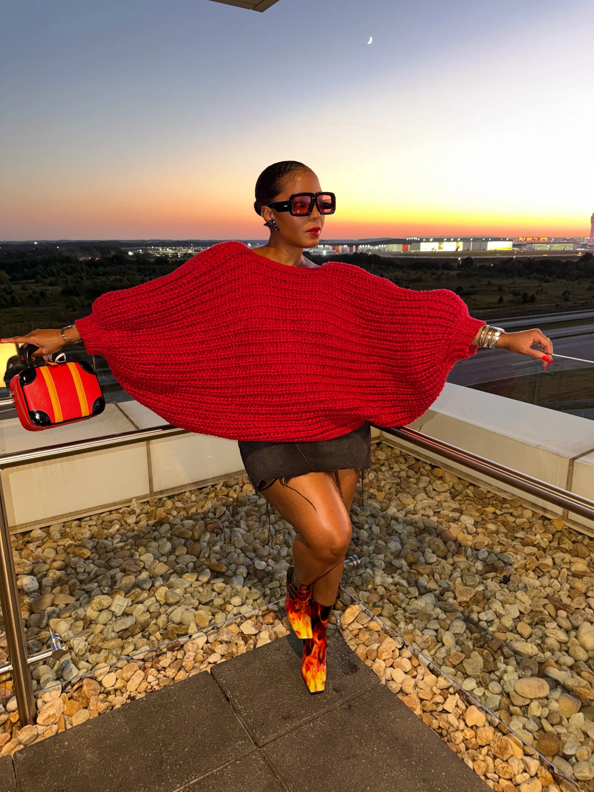 Red oversized knitted sweater with a black mini skirt and tall flame-patterned boots and dark sunglasses.