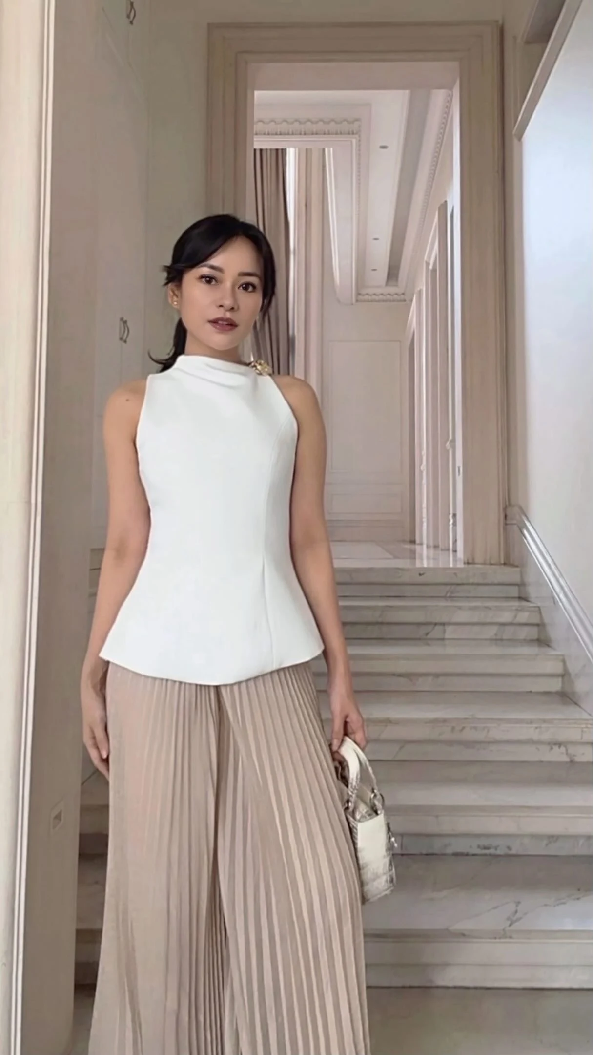 Model on stone stairs in a white sleeveless top and light beige wide-leg pleated trousers with a pale bag.