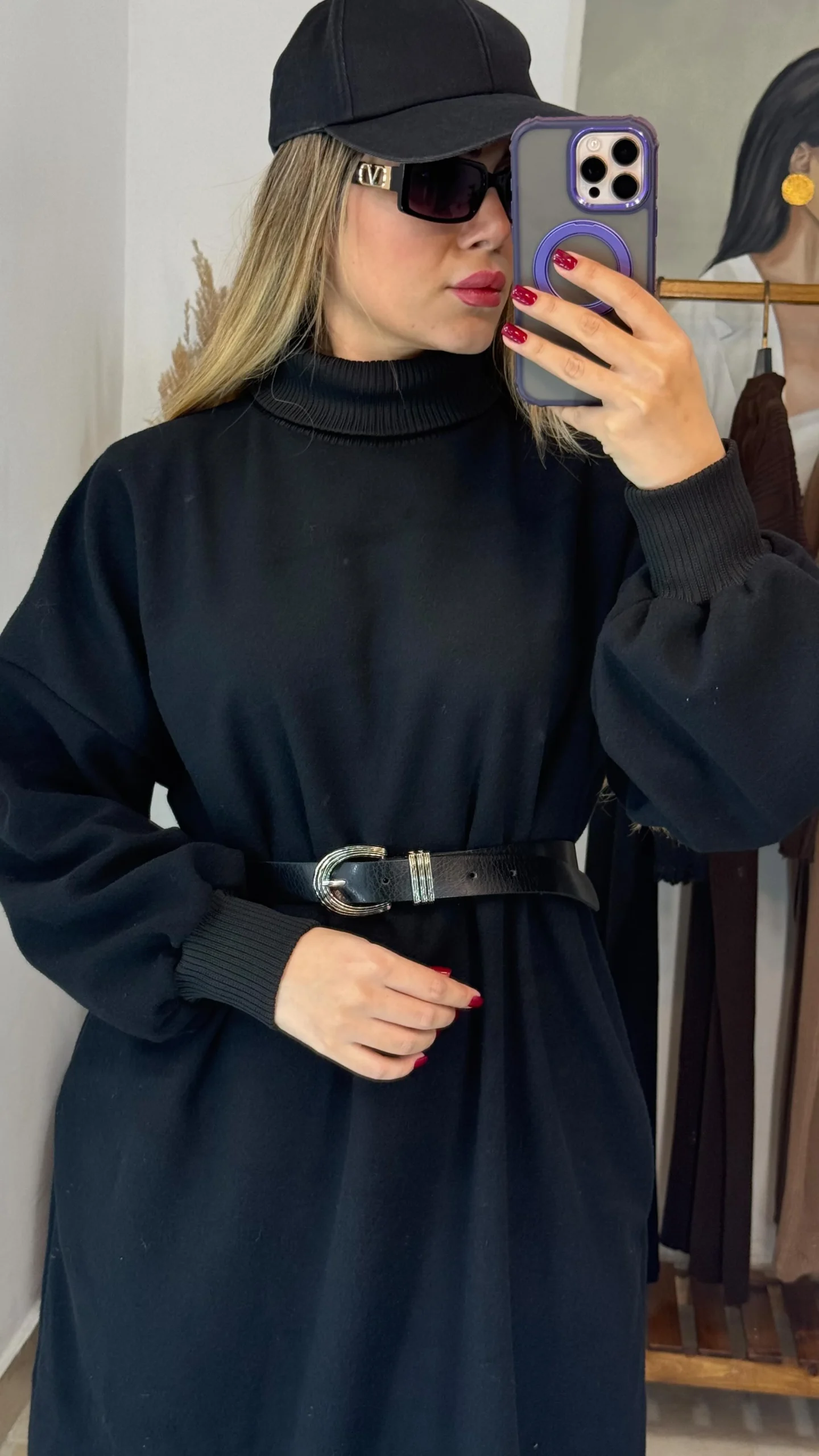 Person in a belted black turtleneck dress, baseball cap, and oversized sunglasses taking a mirror selfie.