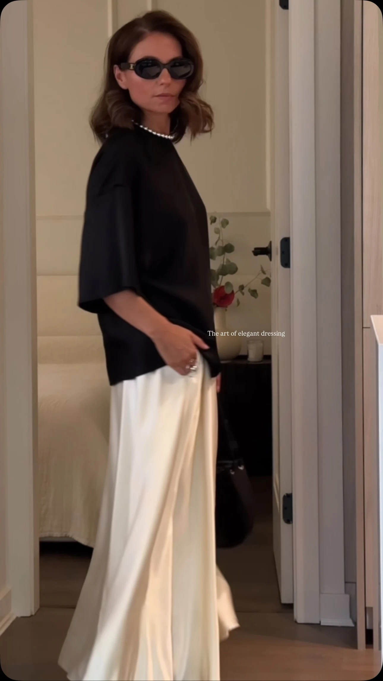 Woman wearing a black top tucked into an ivory satin maxi skirt with pearl necklace and flat sandals.