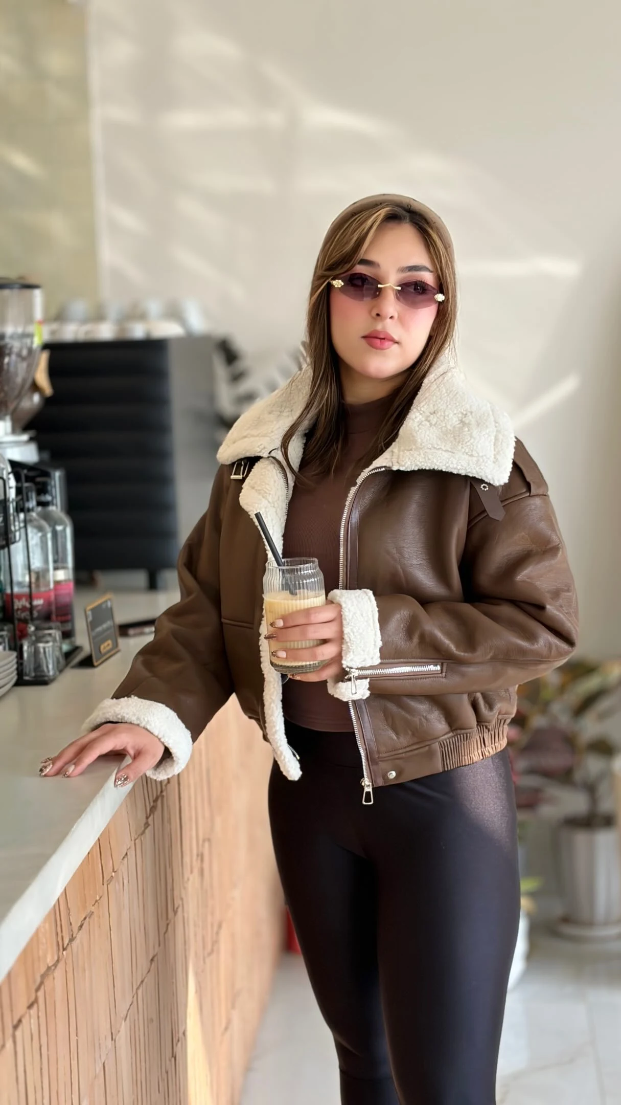 Cropped brown zip jacket with cream collar worn over dark top and fitted black pants.