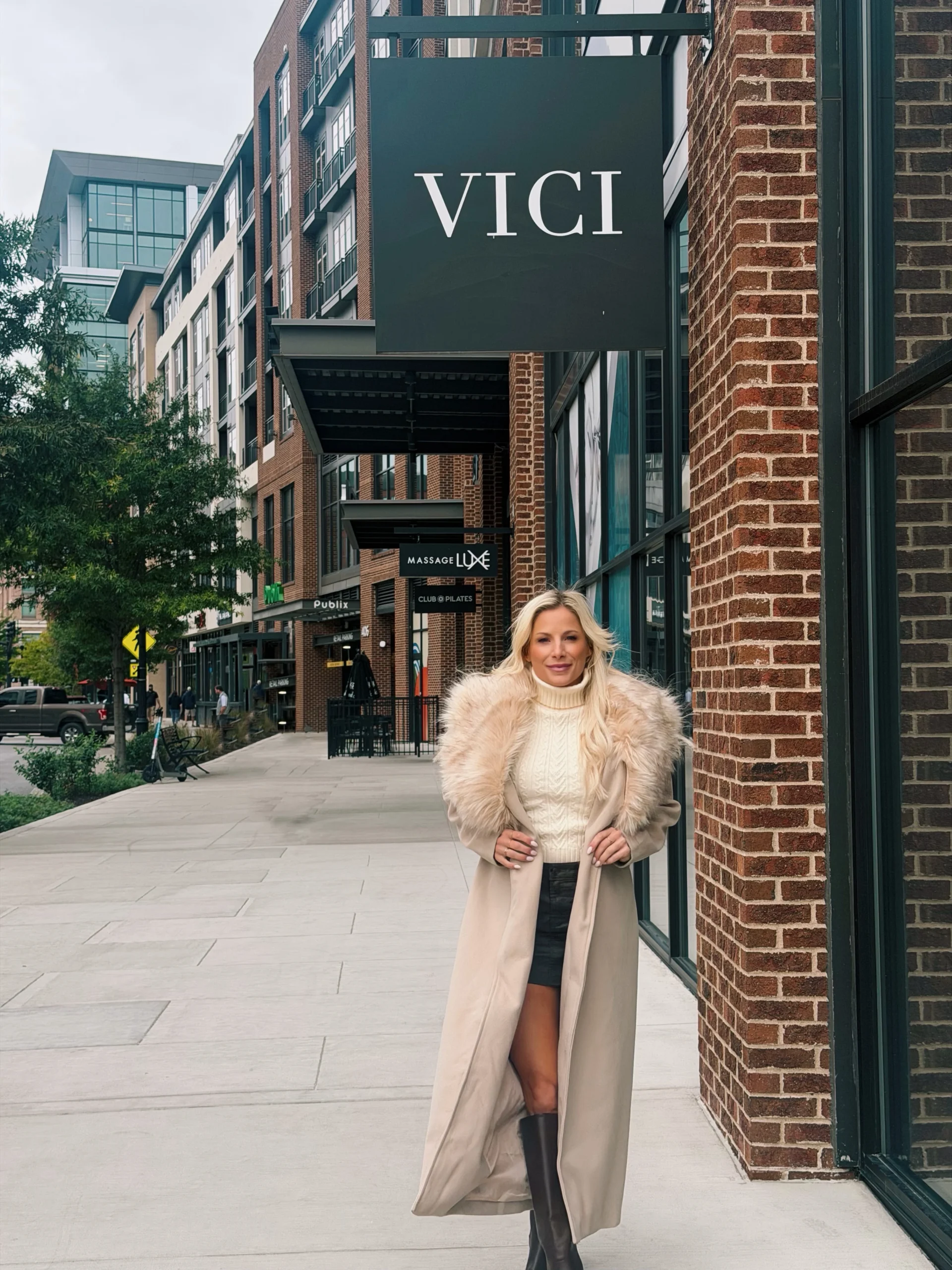 The outfit features a cream cable-knit turtleneck beneath a long, light beige coat with a large fluffy pale collar and matching cuffs. A short dark skirt and tall black boots complete the look. The coat is worn open, showing the sweater and skirt.