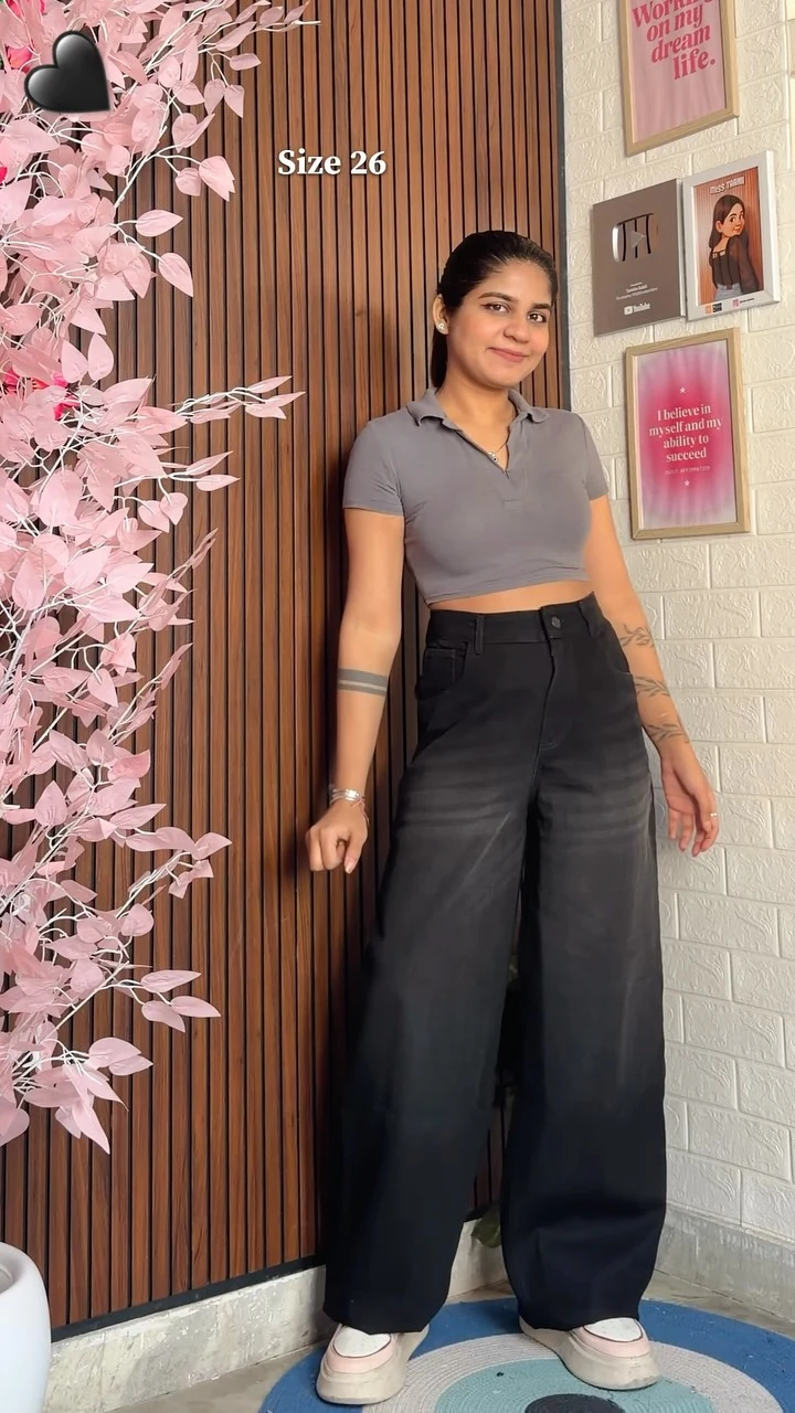 Person in gray cropped polo, black wide-leg jeans, and white sneakers in front of wood-slat wall.
