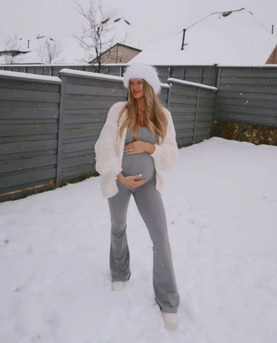 Pregnant woman in gray flared pants, white textured cardigan, and fluffy white hat outdoors in snow.