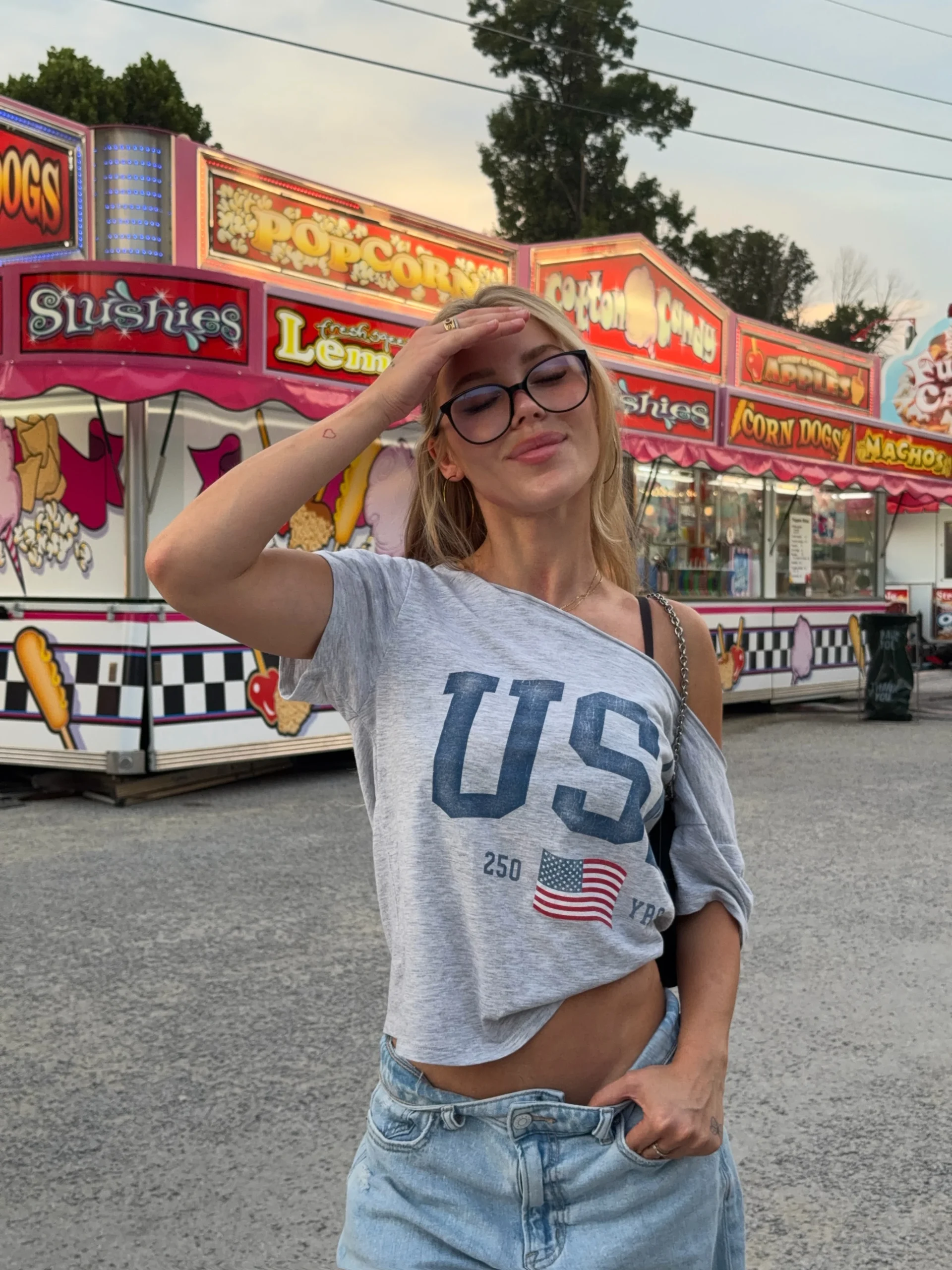 Woman wearing a gray off-the-shoulder USA graphic tee, light-wash baggy jeans, and black crossbody bag.