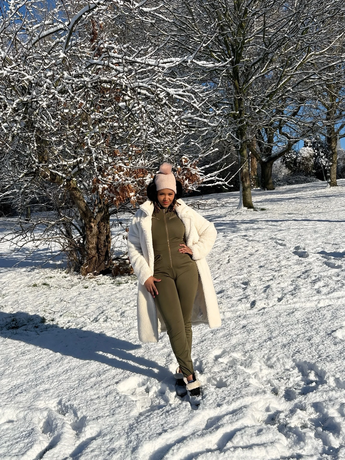 Fitted olive jumpsuit layered under a long white plush coat with a pink pom-pom beanie.