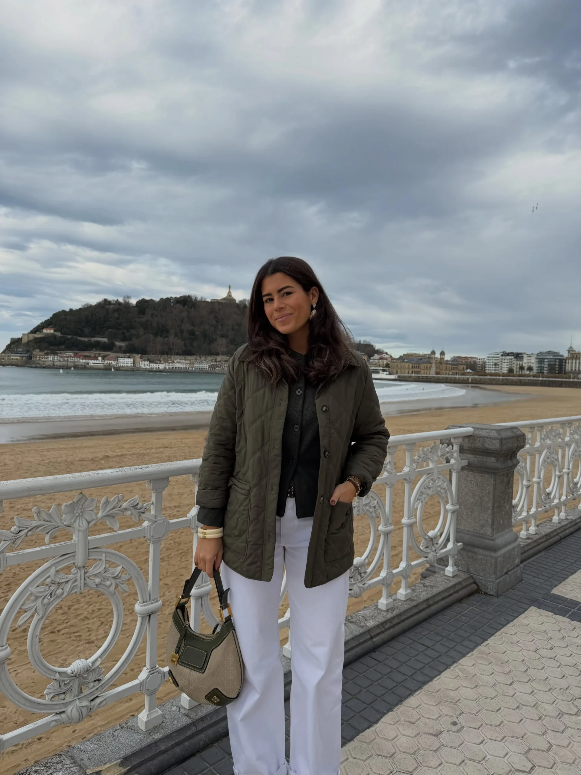 Person in olive quilted jacket, white trousers, and beige-olive bag at a waterfront promenade.