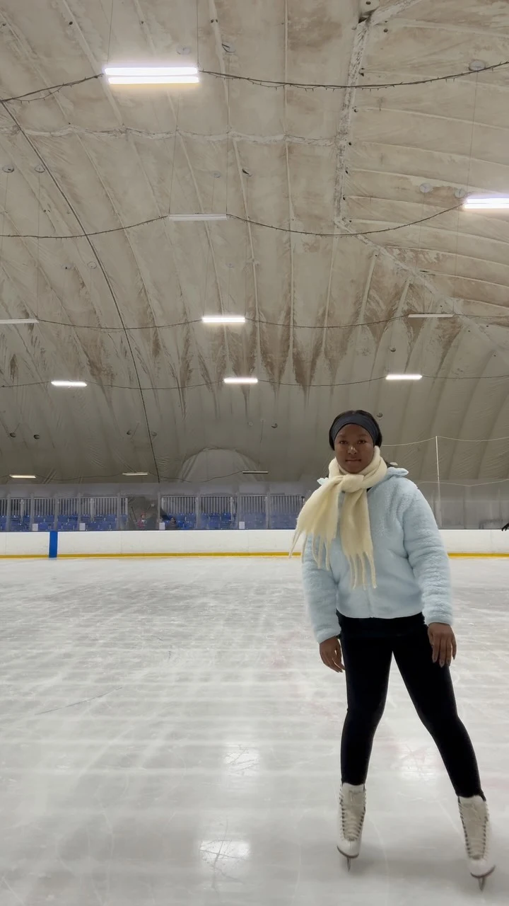 Person on an indoor ice rink in a pale blue hoodie, cream scarf, black leggings, and figure skates.