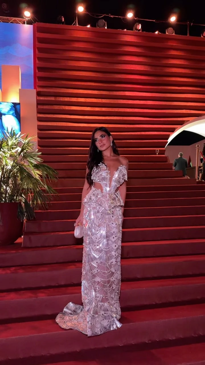 A woman wears a floor-length pale silver off-the-shoulder gown with fan-shaped embellishments and a pale clutch.