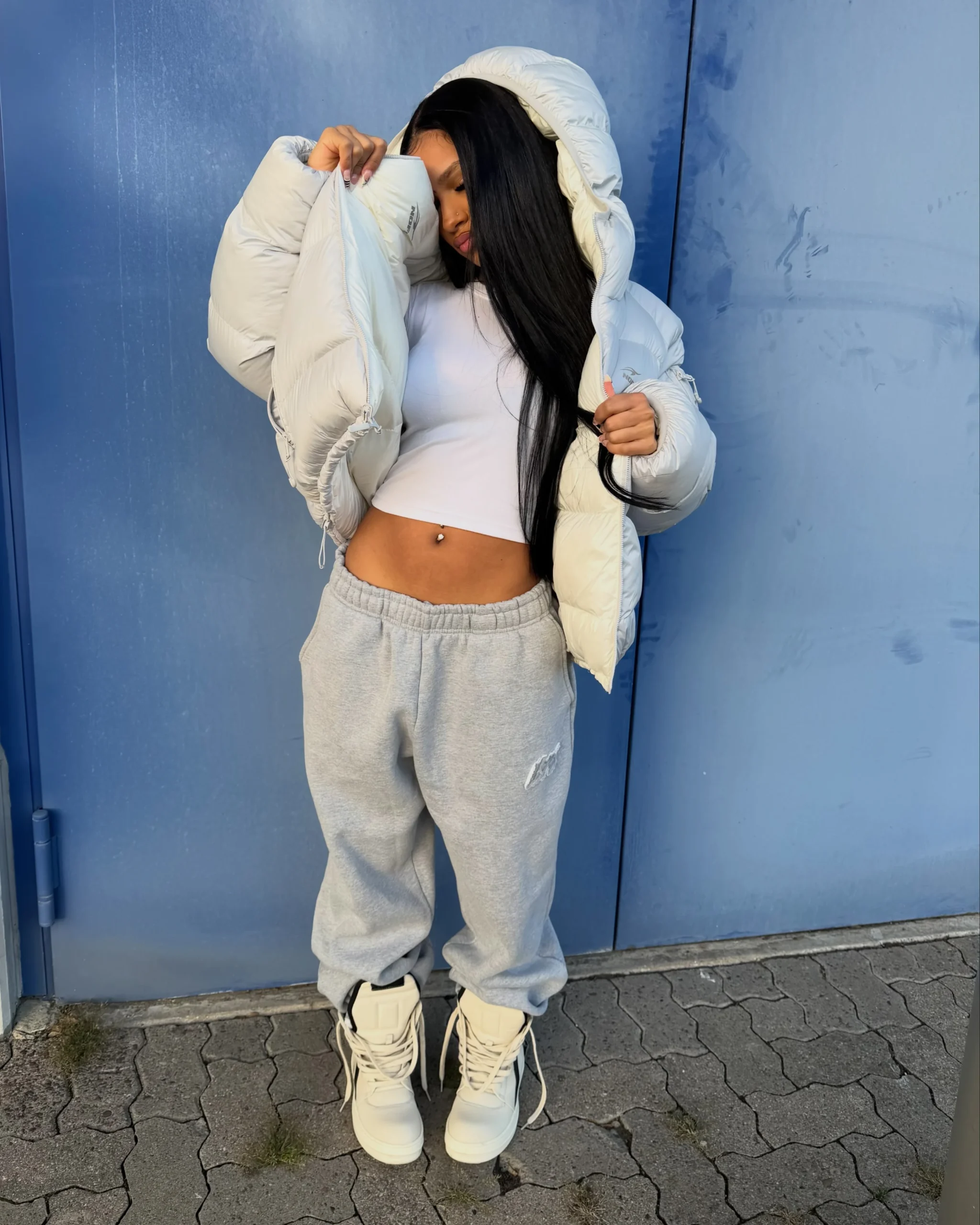 White hooded puffer jacket worn open over a cropped top with gray joggers and cream high-tops.