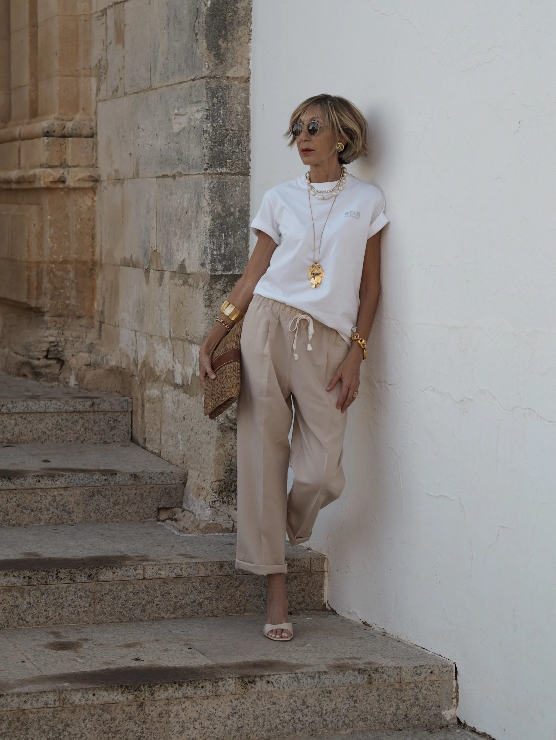 Person in white tee and beige drawstring cropped trousers with gold jewelry and woven clutch.