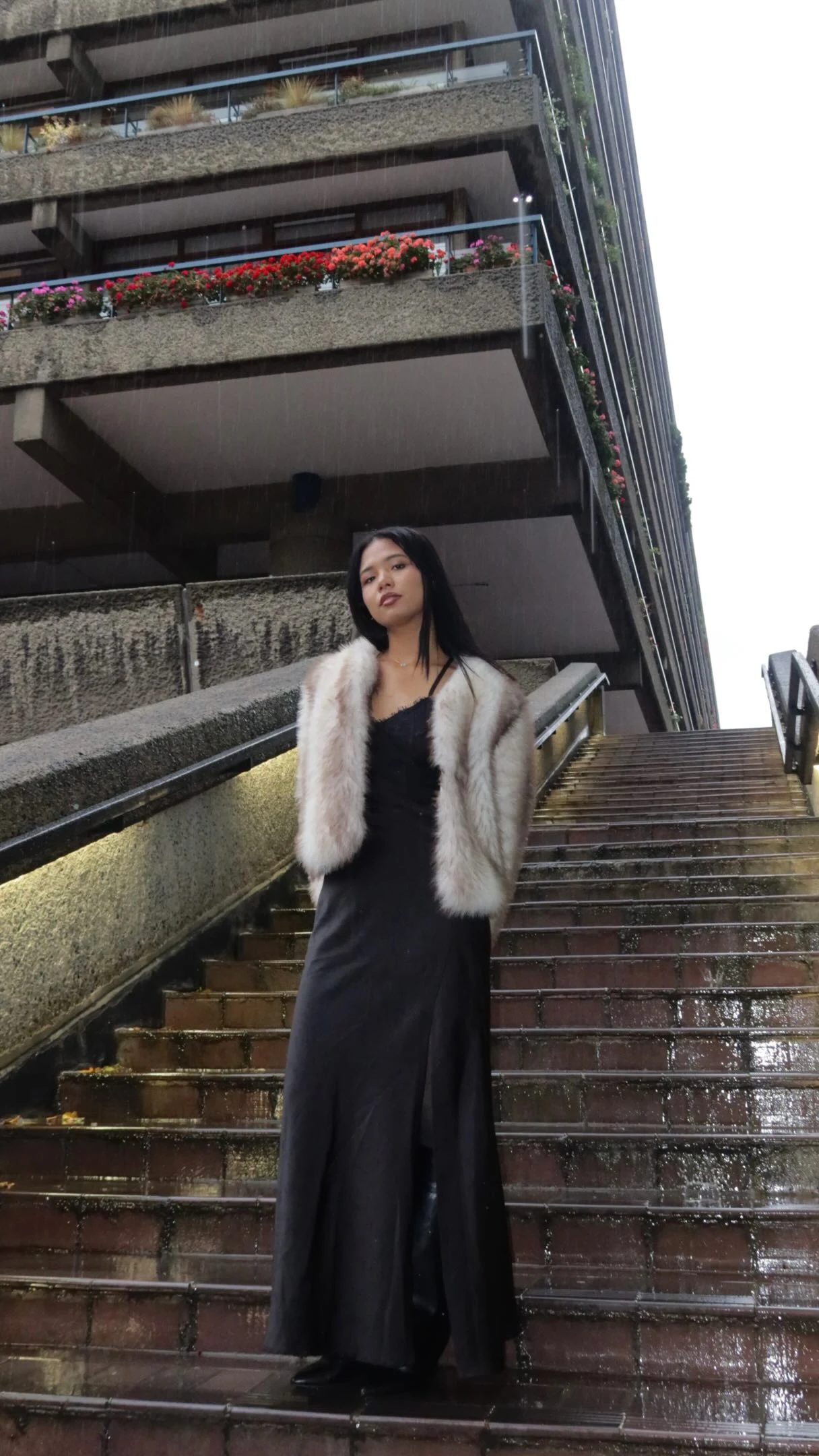 Long black slip dress layered under a short pale cream plush jacket on outdoor stairs.