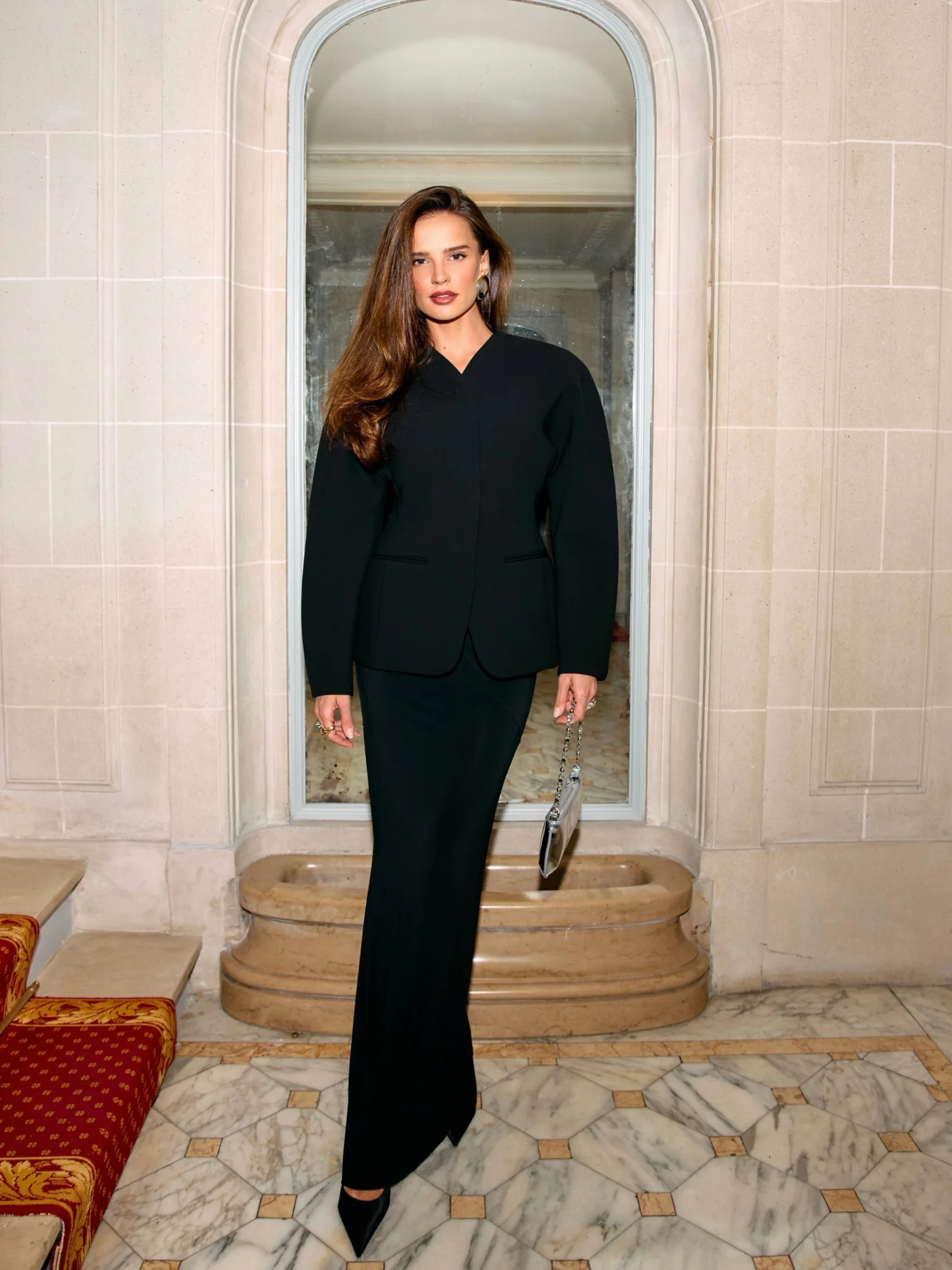 Woman in a black structured blazer, long fitted skirt, pointed heels, and a small silver handbag.