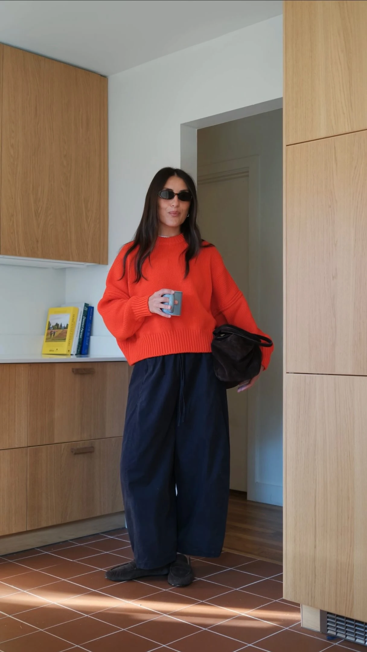 Person wearing a cropped red sweater, wide-leg navy trousers, dark sunglasses, and holding a black bag.