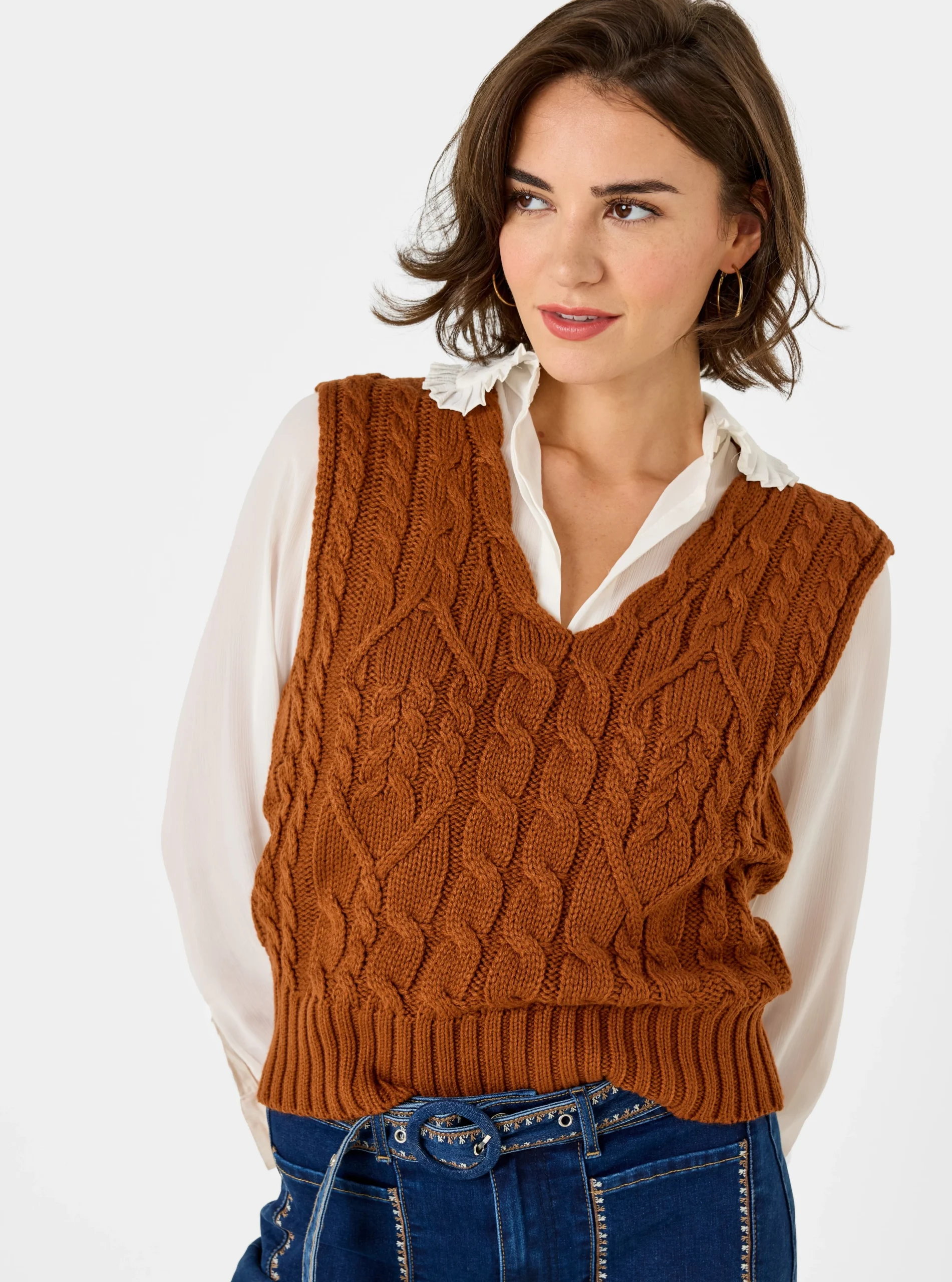 Brown cable-knit sweater vest over a white ruffled collar blouse and dark blue jeans.