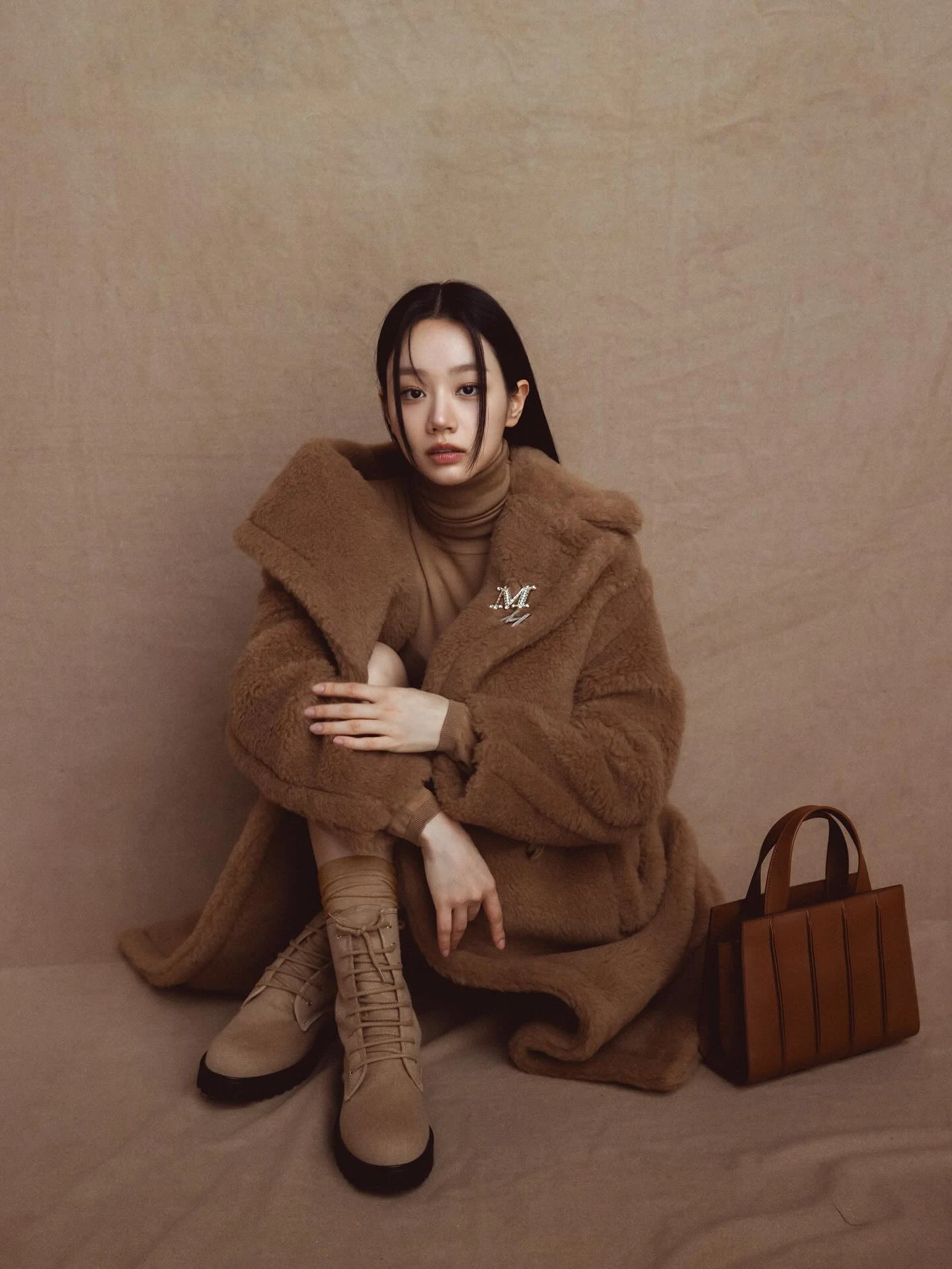 Seated figure in all-brown oversized coat, high-neck top, and lace-up boots beside structured handbag.
