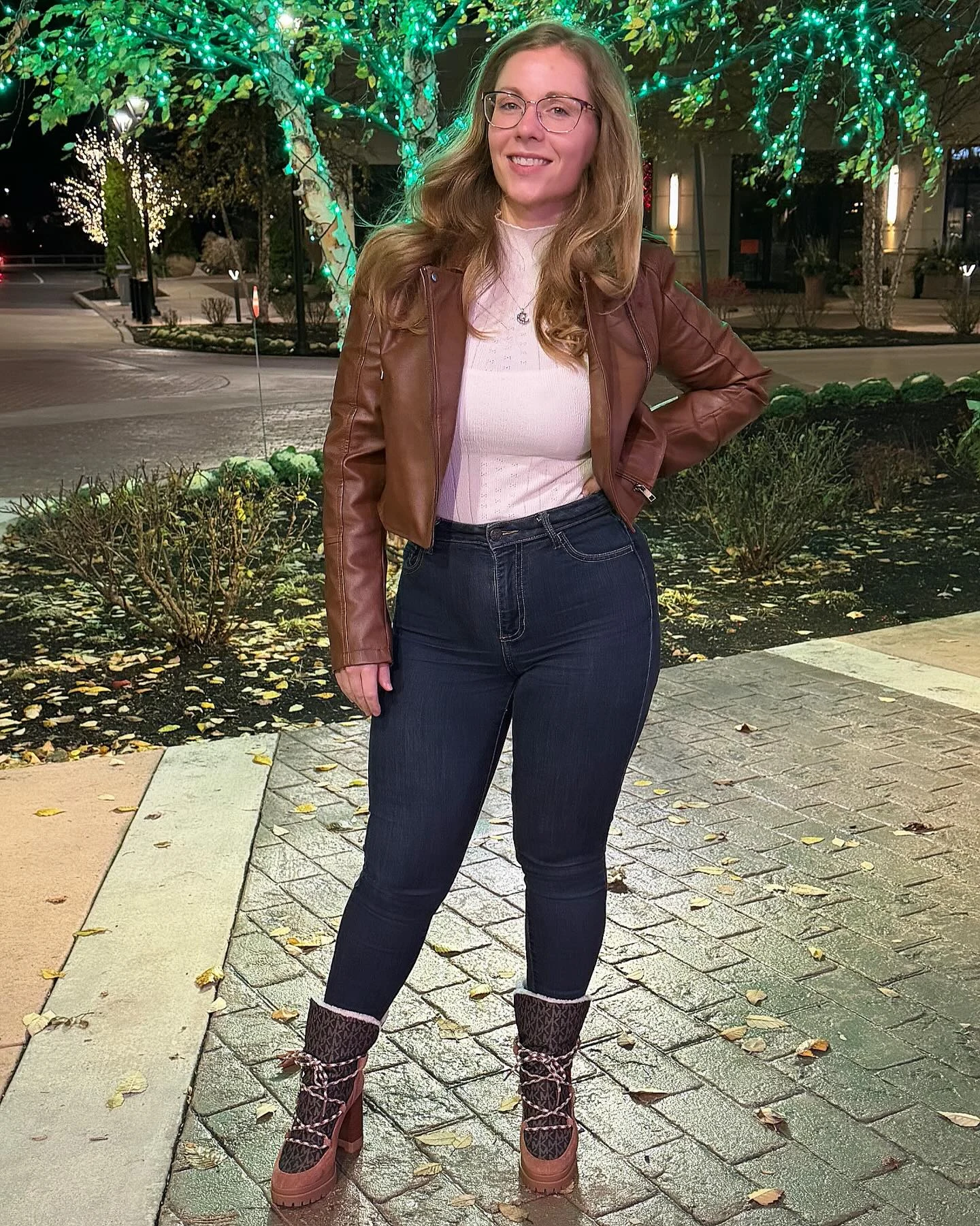 The outfit pairs a cropped brown jacket with a pale pink fitted top and dark, high-waisted skinny jeans. The jeans have turned-up hems. Tall brown boots have chunky soles, block heels, and dark patterned shafts with light-colored laces. A small pendant necklace is visible.