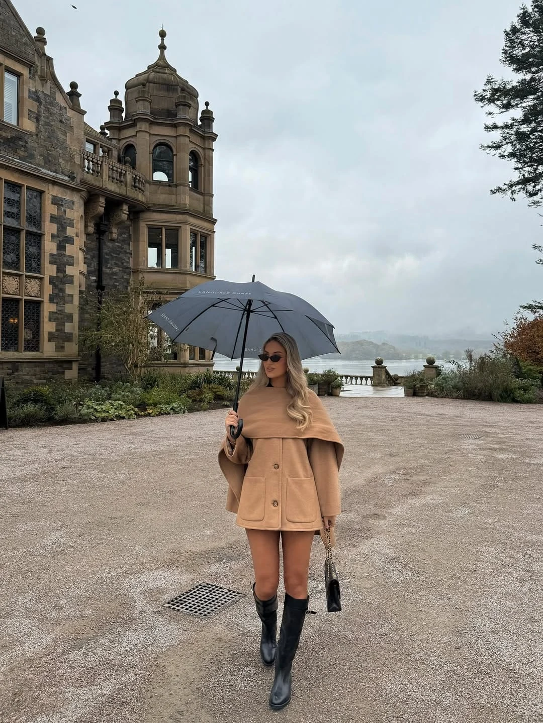 A camel cape coat worn as a dramatic outerwear piece over a mini, with tall black boots and bare legs, is the most editorial interpretation of country chic in this roundup — the cape silhouette has a sweeping, almost architectural quality that photographs beautifully against an outdoor backdrop, and the combination of warm camel and black boots is one of the most graphic and high-contrast pairings in the neutral palette. The sleek umbrella is not a styling afterthought here; it is a prop that makes the whole look feel like a scene rather than just an outfit, giving it a storybook countryside quality that is completely irresistible. I find this completely beautiful for its willingness to be dramatic — country chic does not always have to be understated.