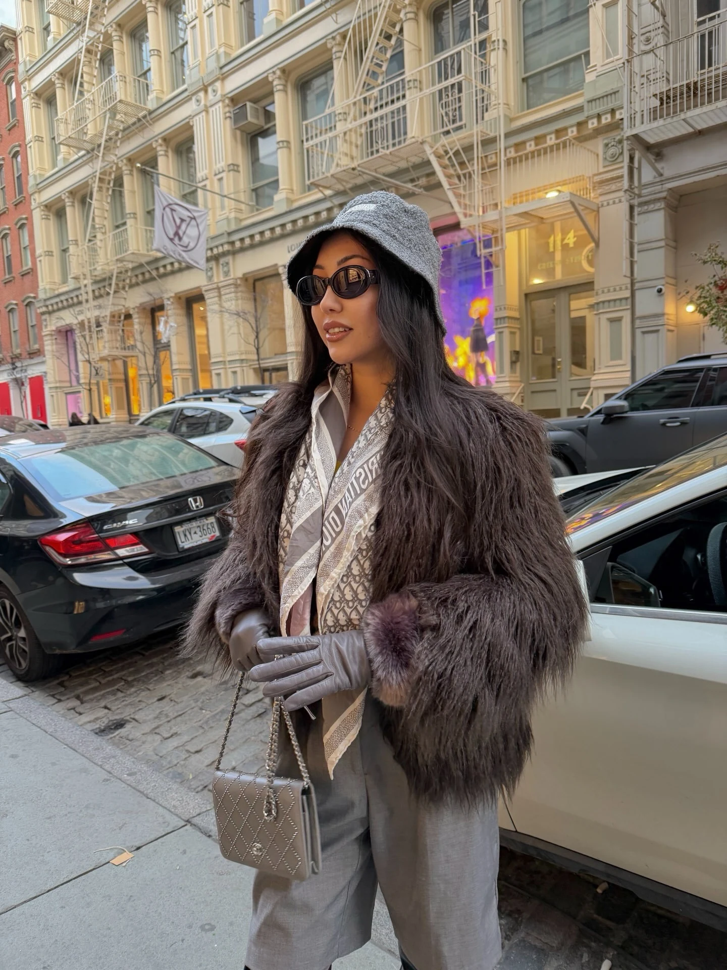 Person in charcoal fur coat, gray brimmed hat, loose gray trousers, and taupe chain-strap bag.