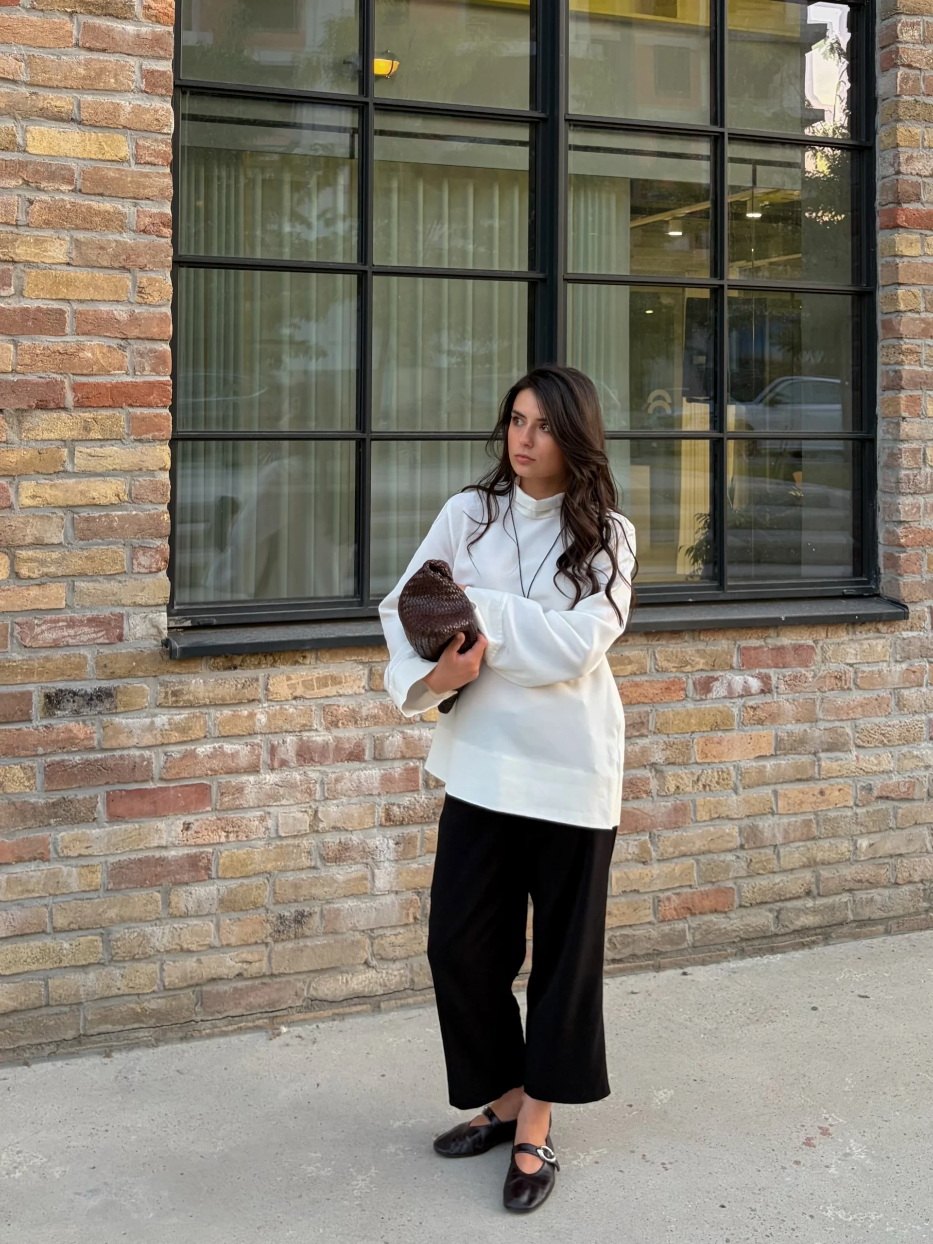 Loose white long-sleeve top, black wide-leg trousers, brown woven clutch, and dark flat shoes on a woman.