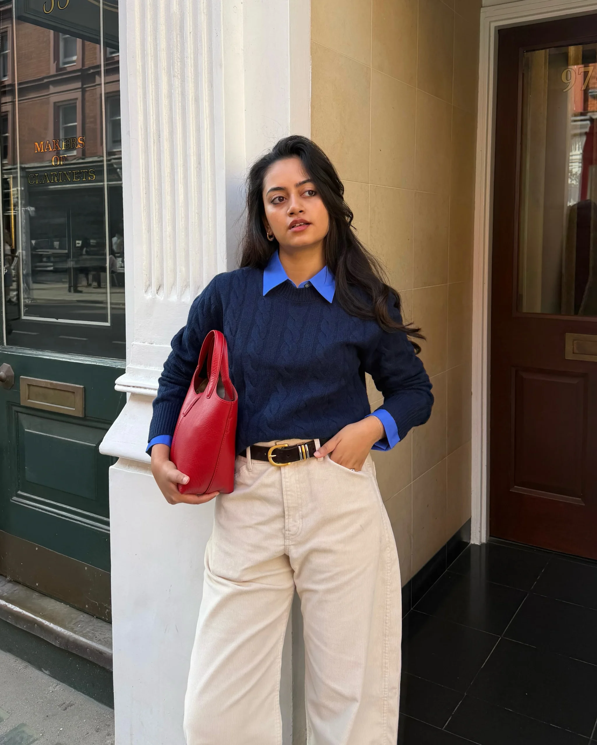 Navy cable knit over a blue shirt, cream wide-leg trousers, brown belt, black boots, and red tote.